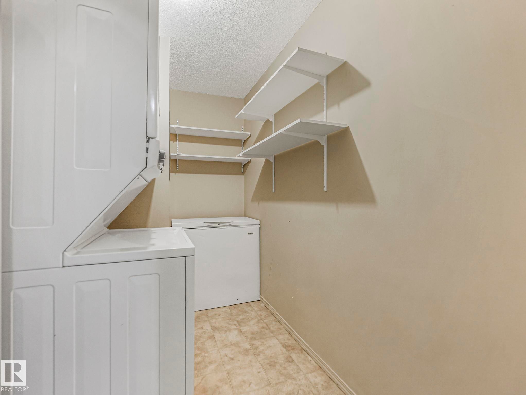 Dedicated laundry area featuring a stacked washer and dryer unit, multiple wall-mounted shelves, and neutral-toned flooring - #143 10636 120 St Nw, Edmonton, AB - Indoor Photo Showing Laundry Room
