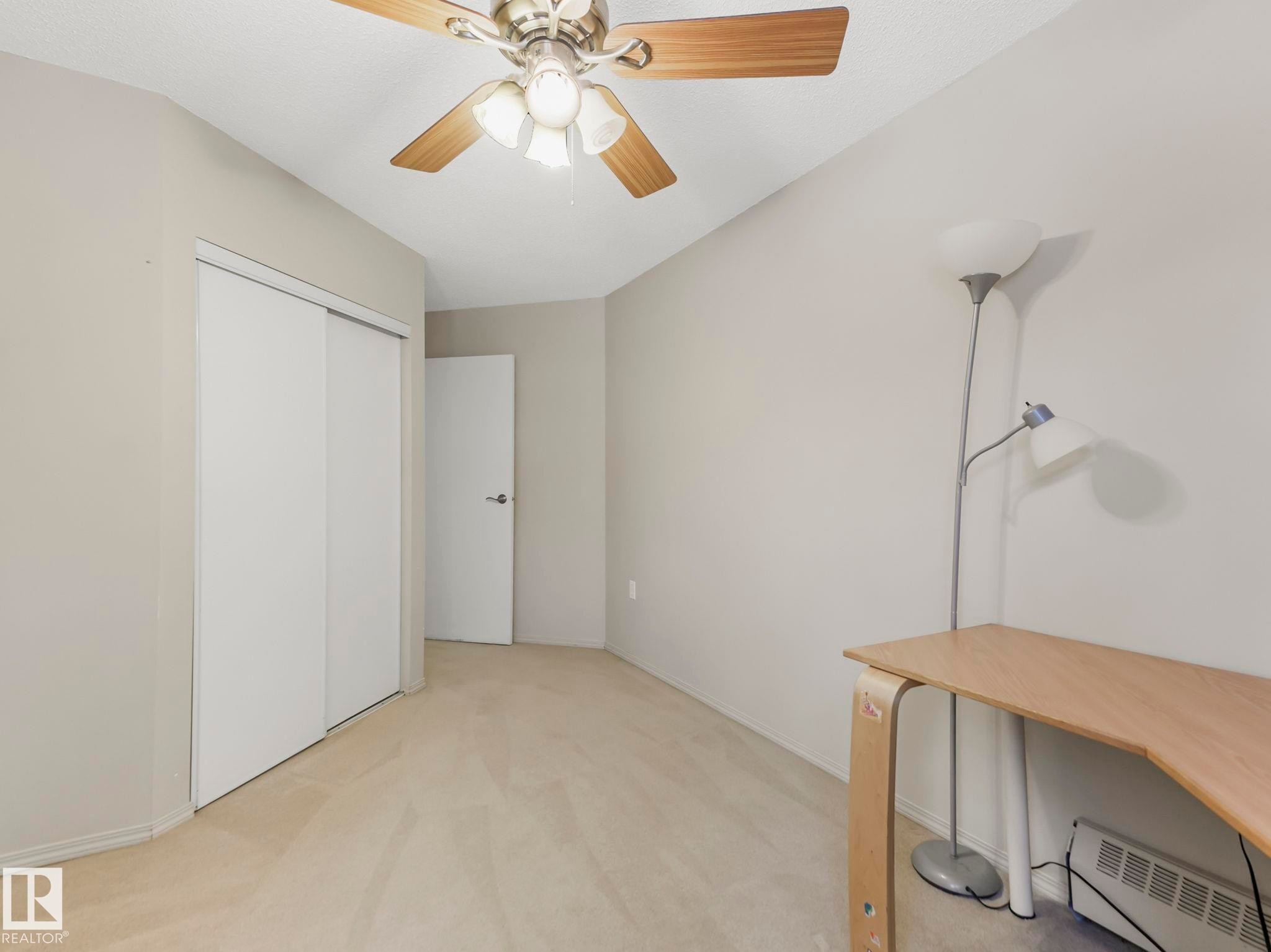 Light-toned carpeting complements neutral wall paint in this room featuring a ceiling fan with wood-finish blades, white sliding closet doors, and a white interior door - #143 10636 120 St Nw, Edmonton, AB - Indoor Photo Showing Other Room