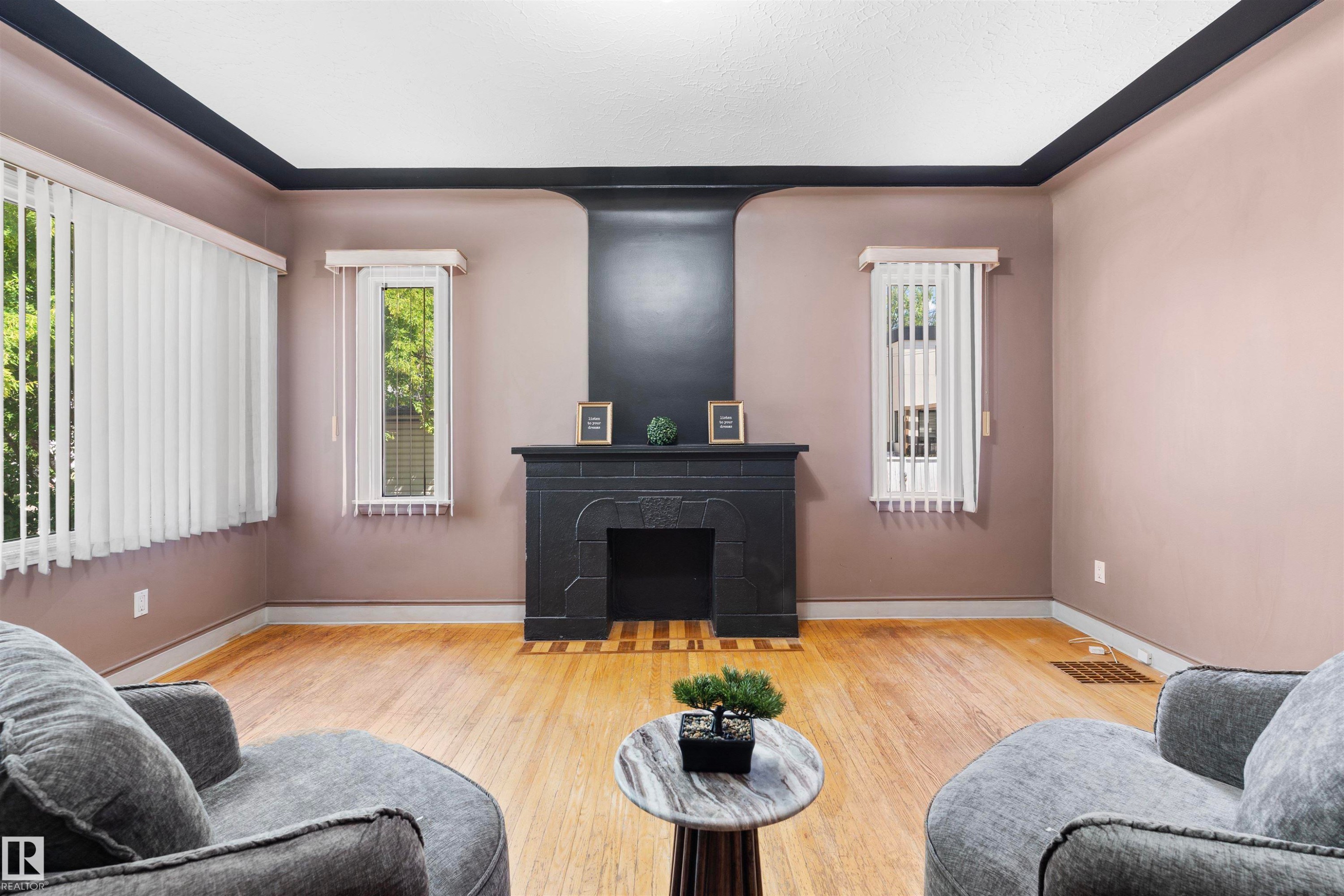 Living area featuring a black fireplace with a mantel, light wood-finish flooring, two large windows with vertical blinds, and dark ceiling trim - 10607 73 Ave, Edmonton, AB