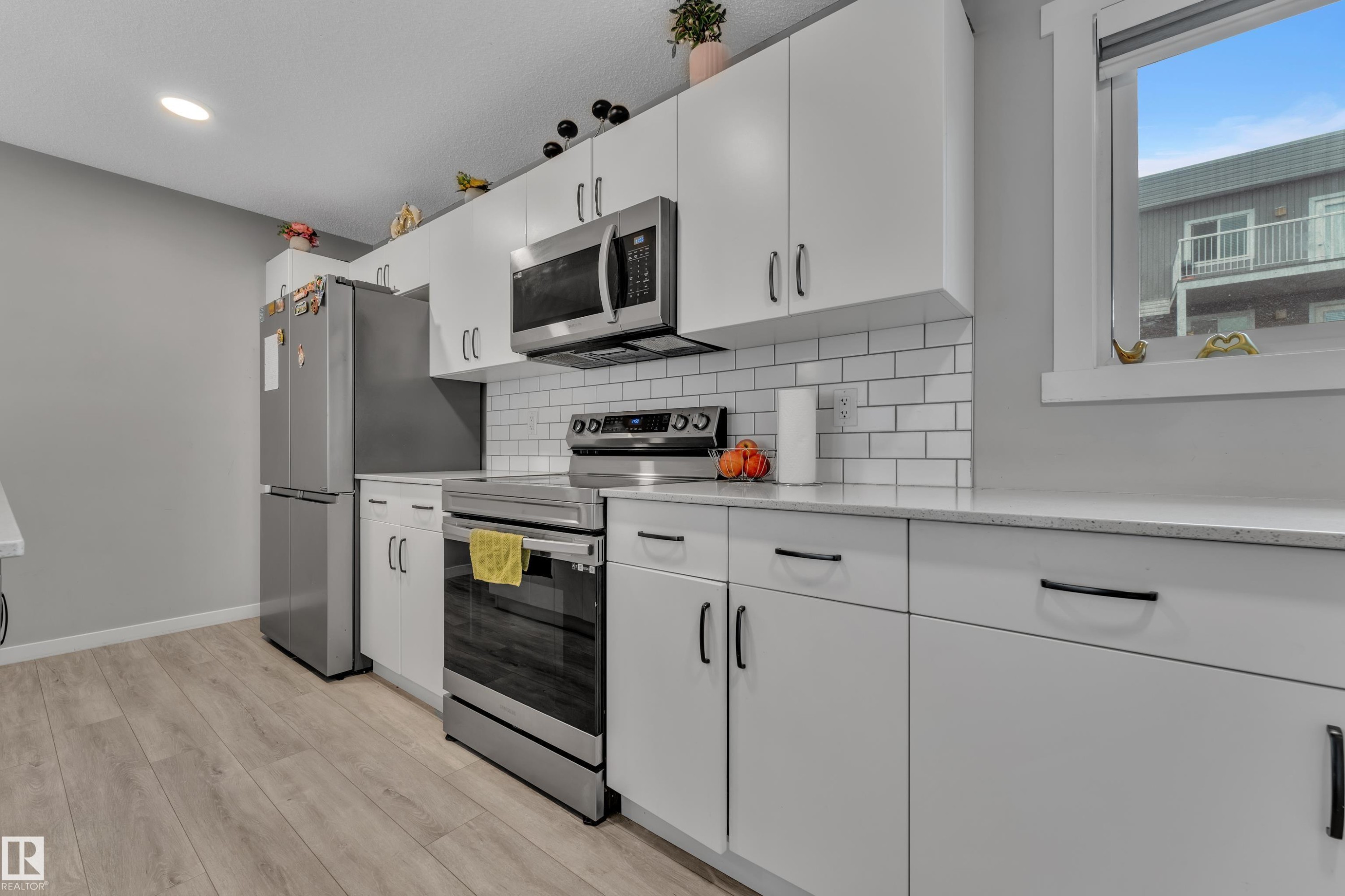Contemporary kitchen featuring white cabinetry with black hardware, stainless steel appliances, a white subway tile backsplash, light-toned countertops, and wood-finish flooring - #82 1816 Rutherford Rd Sw, Edmonton, AB - Indoor Photo Showing Kitchen