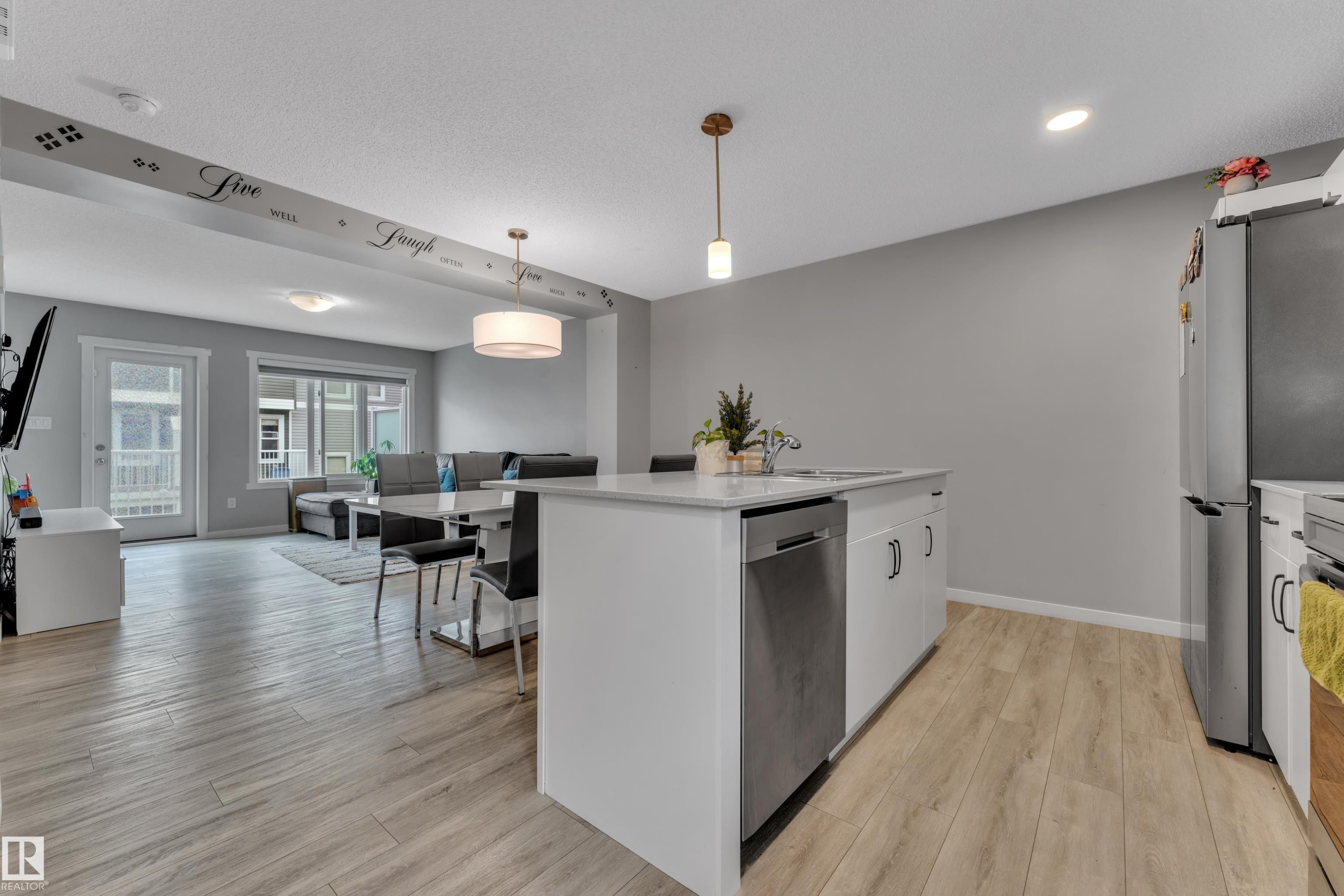 Open-concept living space featuring wood-finish flooring, a kitchen island with integrated sink, stainless steel dishwasher, and a contemporary pendant light fixture - #82 1816 Rutherford Rd Sw, Edmonton, AB - Indoor Photo Showing Kitchen With Upgraded Kitchen