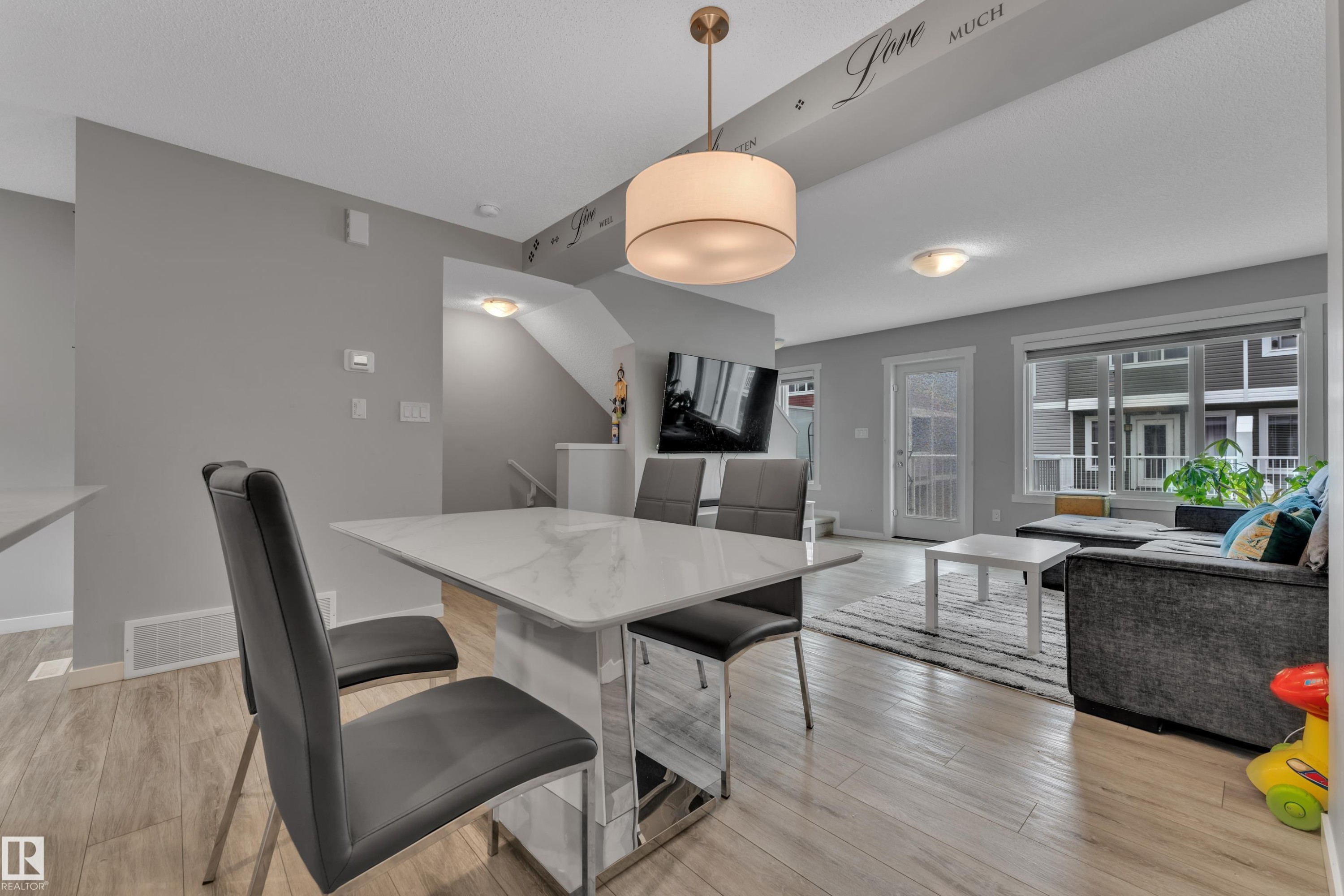 Open-concept living area featuring light wood-finish flooring, a contemporary drum pendant light, and abundant natural light from a large window - #82 1816 Rutherford Rd Sw, Edmonton, AB - Indoor