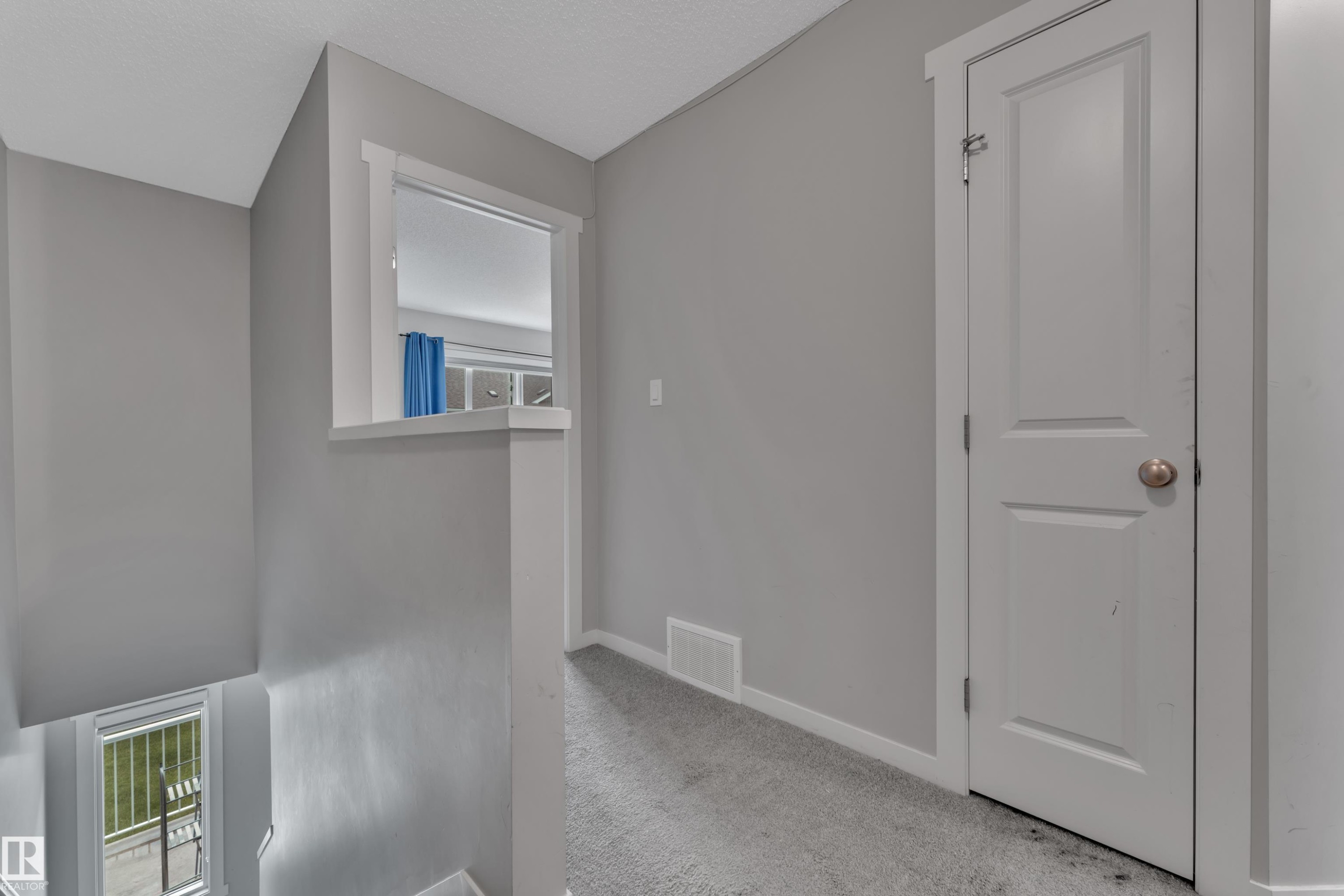 Carpeted hallway featuring light gray walls, white trim, a solid white door with a round brass-tone knob, and an architectural half-wall with an interior window - #82 1816 Rutherford Rd Sw, Edmonton, AB - Indoor Photo Showing Other Room