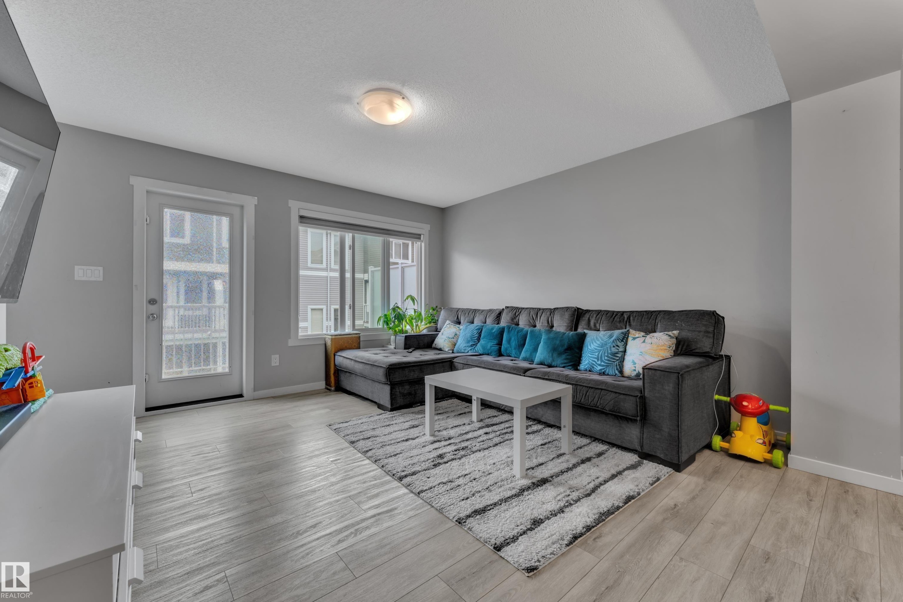 Living area featuring wood-finish flooring, light grey wall tones, and a glass-paneled exterior door - #82 1816 Rutherford Rd Sw, Edmonton, AB - Indoor Photo Showing Living Room