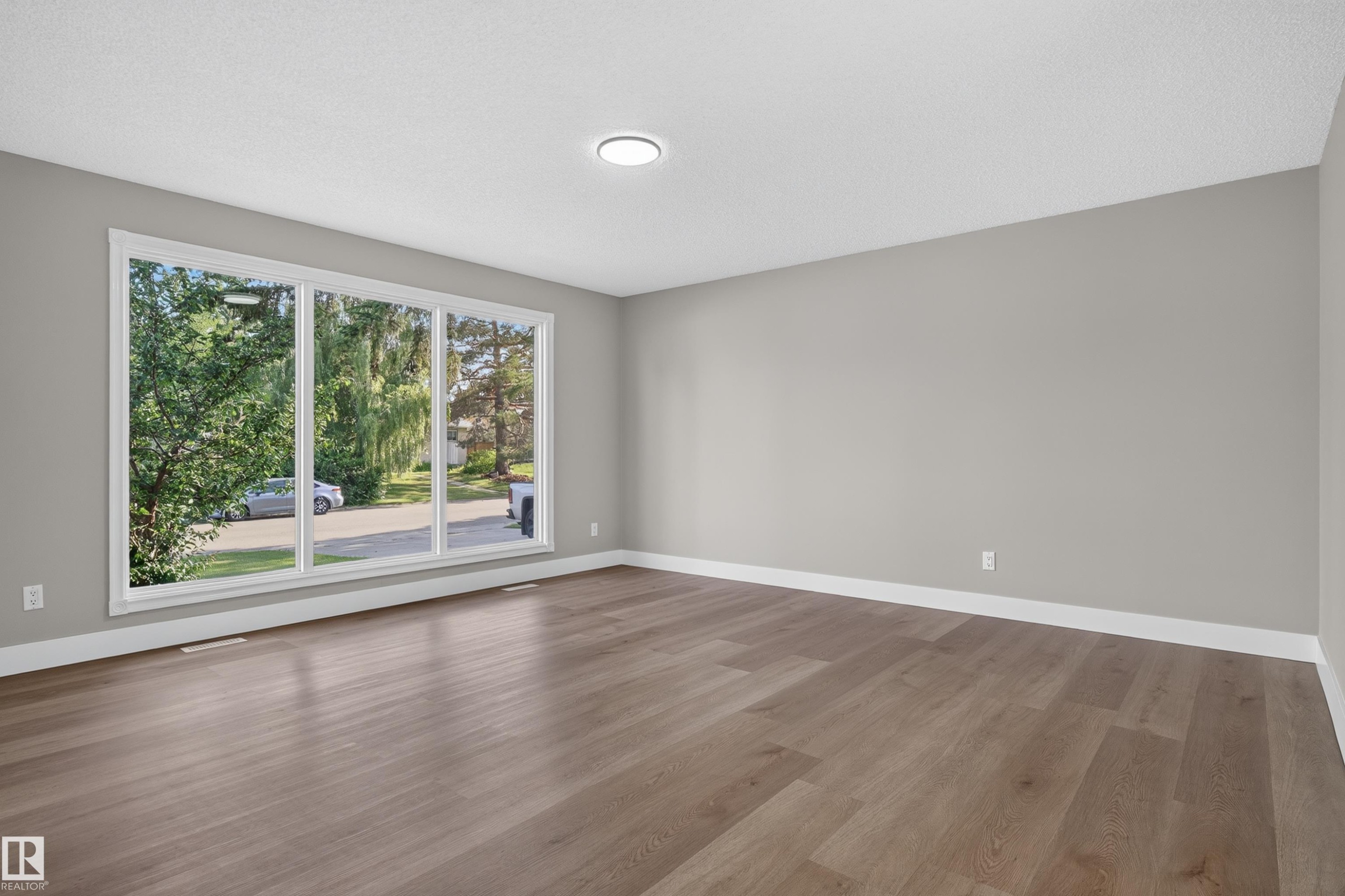Spacious room featuring a large multi-pane window, wood-finish flooring, white baseboards, and a flush-mount ceiling light fixture - 33 Brookside Avenue, Spruce Grove, AB