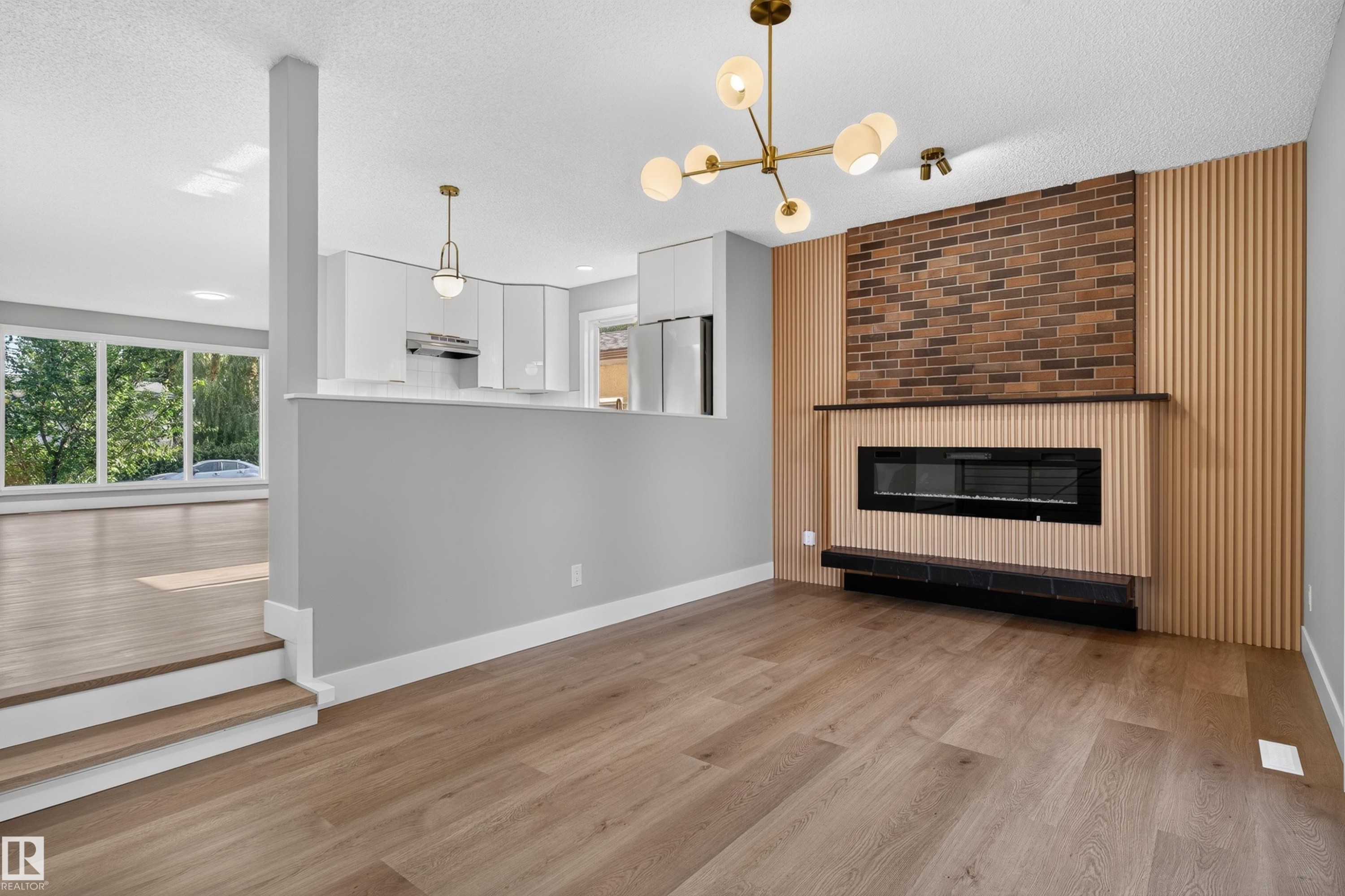 Spacious interior featuring wide plank wood-finish flooring, a modern linear electric fireplace with a brick and wood slat surround, and a contemporary chandelier - 33 Brookside Avenue, Spruce Grove, AB