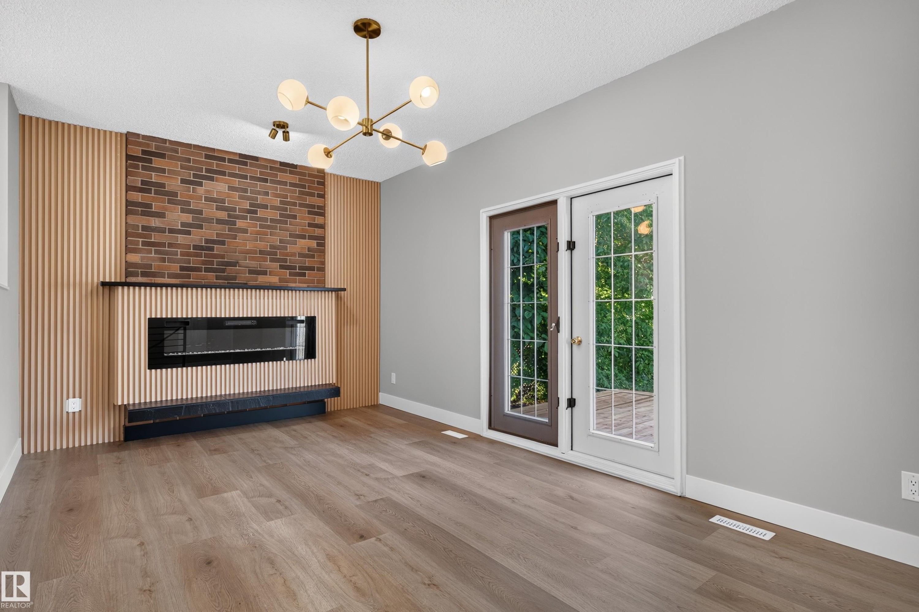 Contemporary living area featuring a wood-panel accent wall with exposed brick, an integrated linear electric fireplace, and a modern brass chandelier - 33 Brookside Avenue, Spruce Grove, AB
