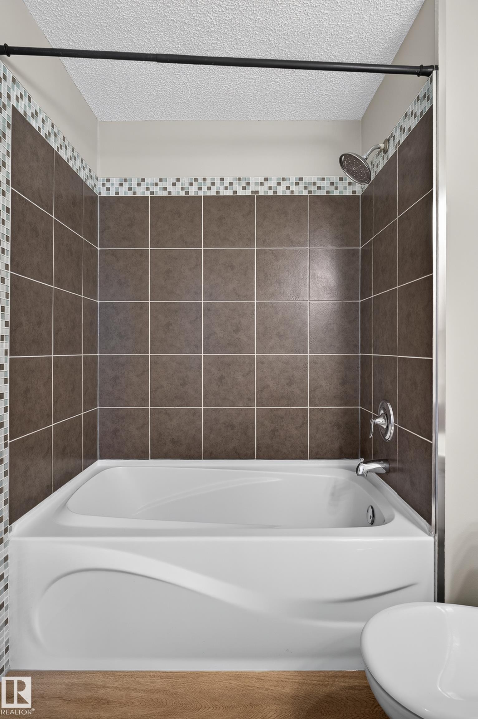 Shower-over-tub combination featuring a white acrylic tub with a curved apron design, brown square wall tiling, a mosaic accent band, and chrome fixtures - 33 Brookside Avenue, Spruce Grove, AB