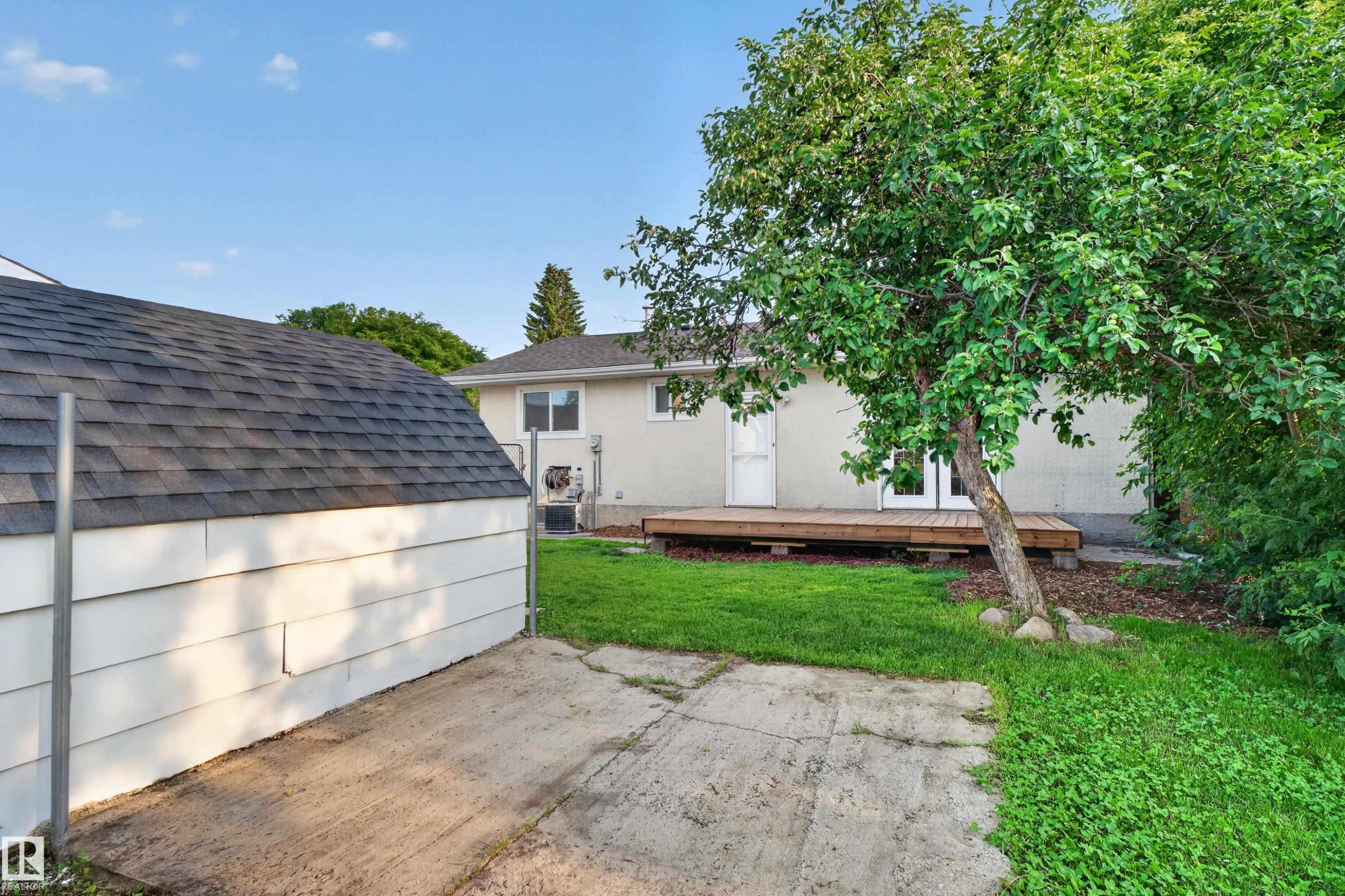 Backyard featuring a wooden deck with double doors, mature trees, a manicured lawn, and a detached shed with shingle roofing - 33 Brookside Avenue, Spruce Grove, AB