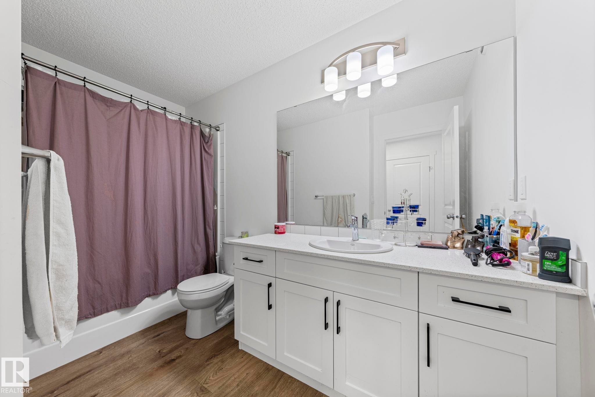 Bathroom featuring a wide vanity with a white countertop and integrated sink, white cabinetry with dark hardware, a large wall-mounted mirror, and wood-finish flooring - 16 19904 31 Avenue, Edmonton, AB