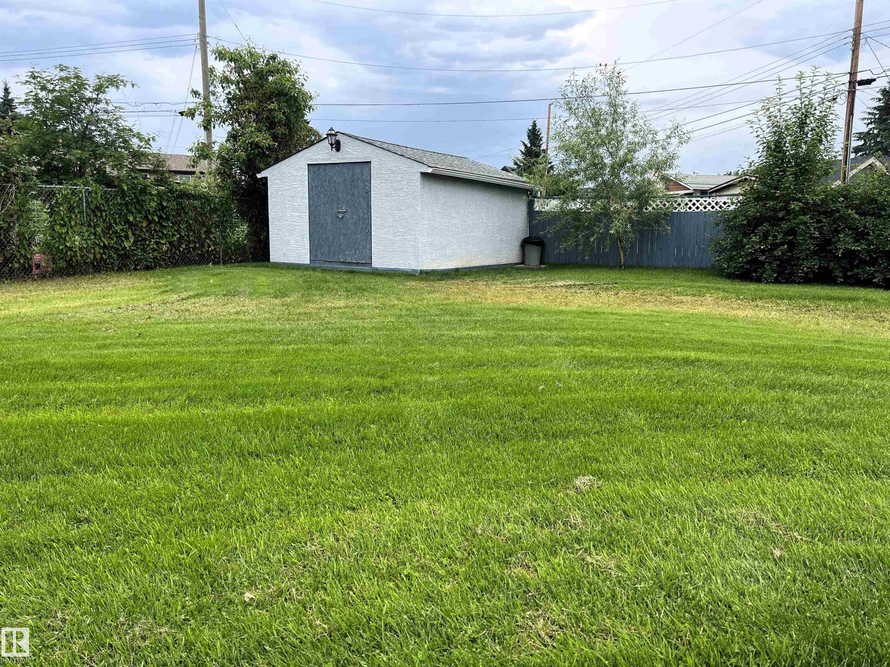 Spacious green lawn with a white storage shed featuring a gray door, mature trees, and perimeter fencing - 4914 51 Ave, Entwistle, AB