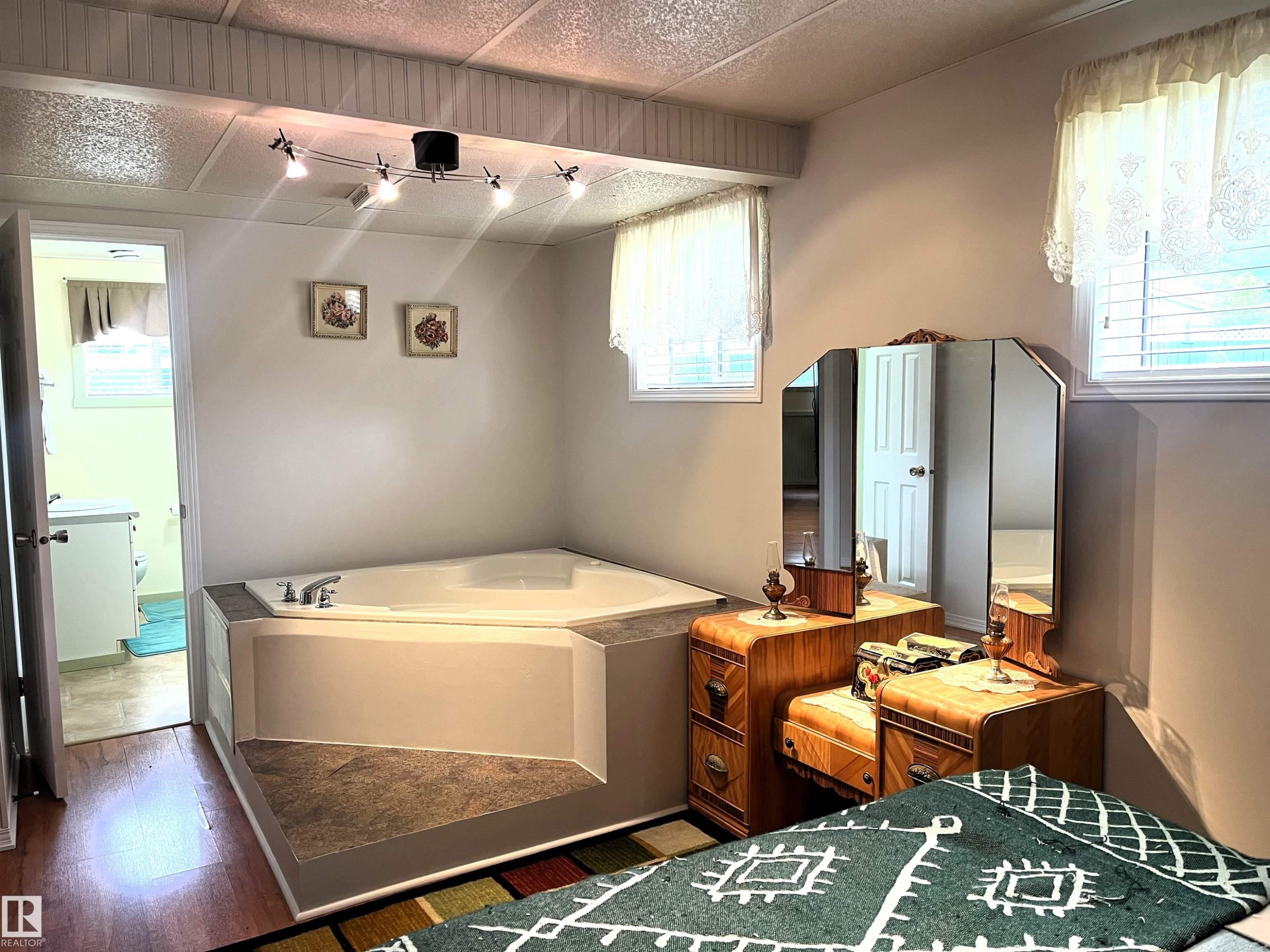 Built-in corner soaking tub with surround, track lighting fixture, two windows with blinds, and wood-finish flooring - 4914 51 Ave, Entwistle, AB
