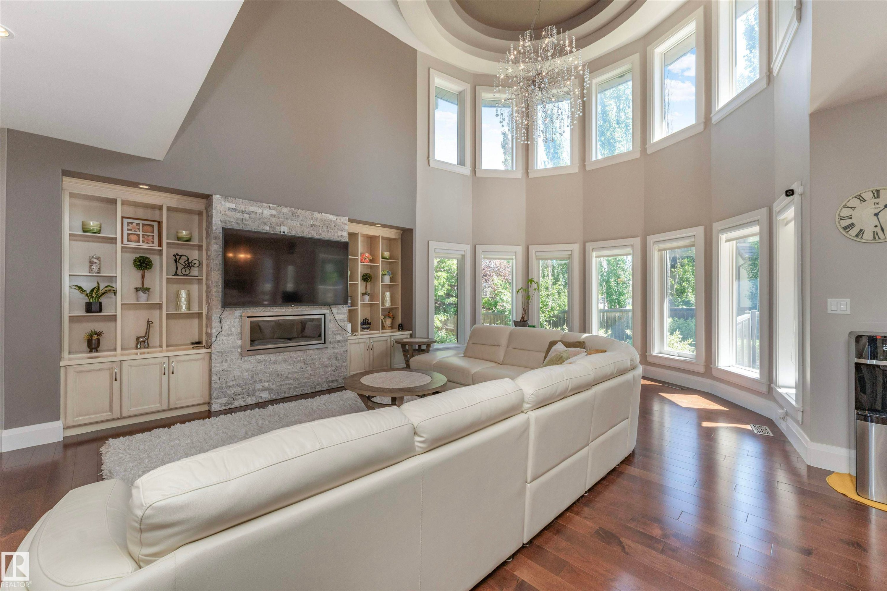 Two-story living area featuring a curved wall of windows, a grand chandelier, hardwood flooring, a stone-clad fireplace, and built-in shelving with cabinetry - 3503 Watson Point(E), Edmonton, AB