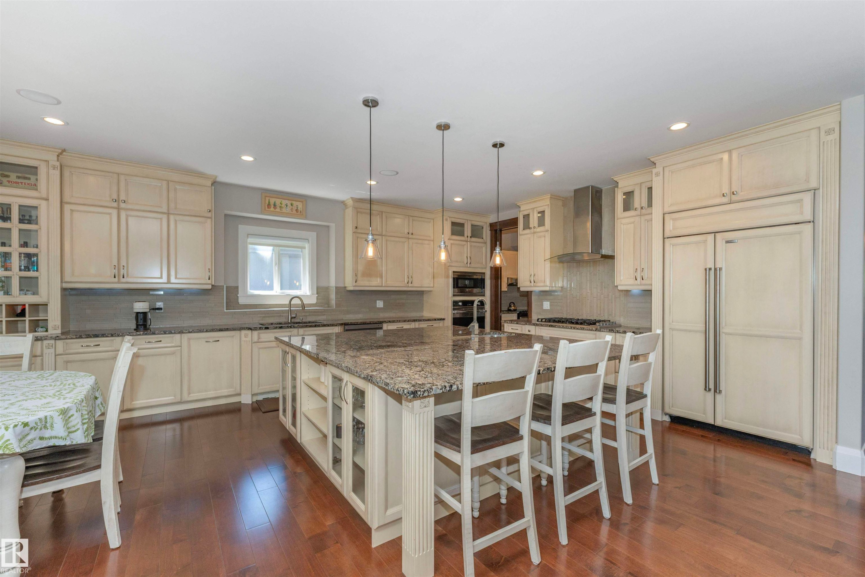 Expansive kitchen featuring a large central island with seating, cream-colored cabinetry, granite countertops, stainless steel appliances, and wood-finish flooring - 3503 Watson Point(E), Edmonton, AB