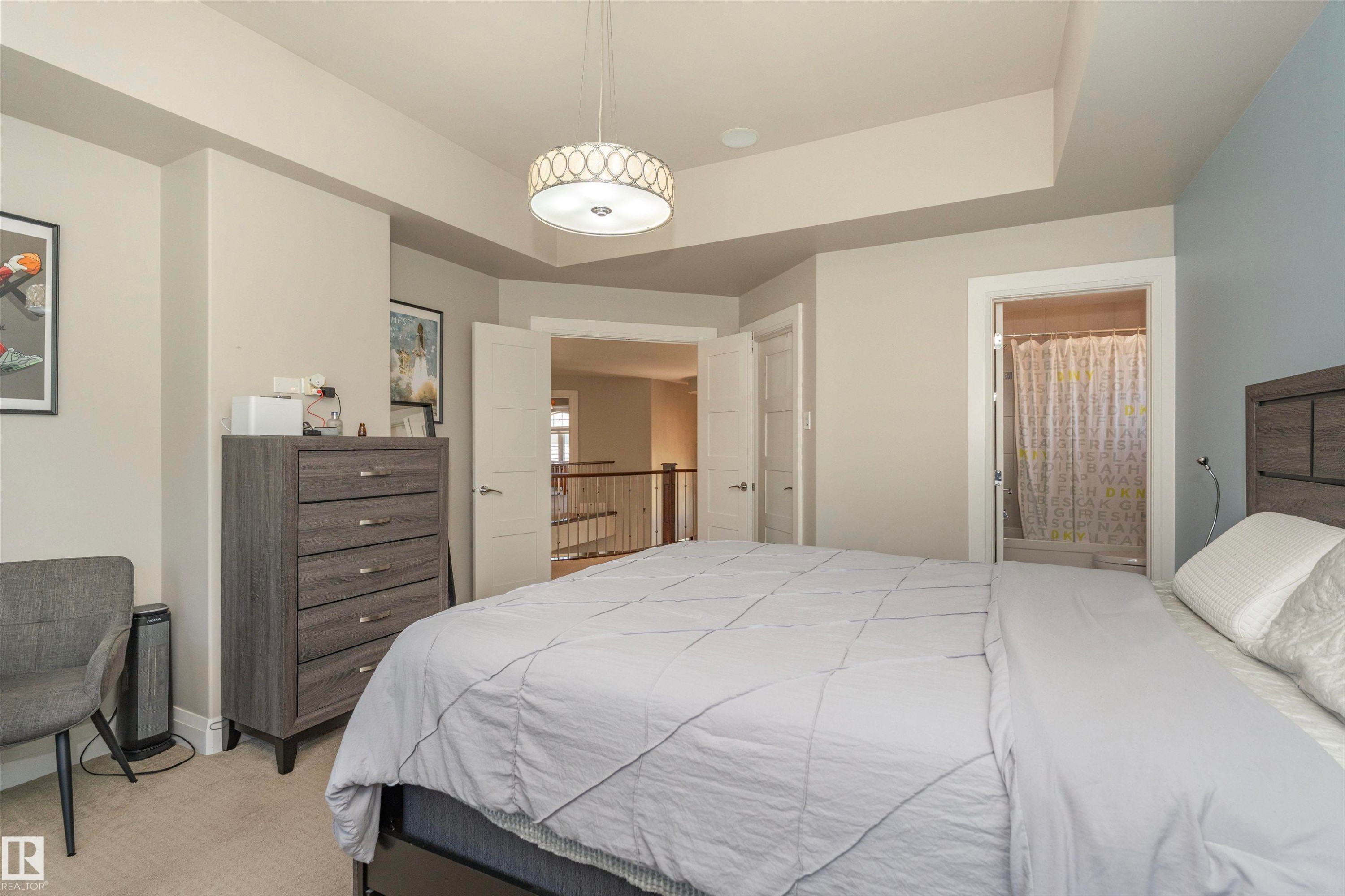 Carpeted bedroom featuring a decorative ceiling light fixture, tray ceiling, and an open doorway leading to a hallway with railings - 3503 Watson Point(E), Edmonton, AB