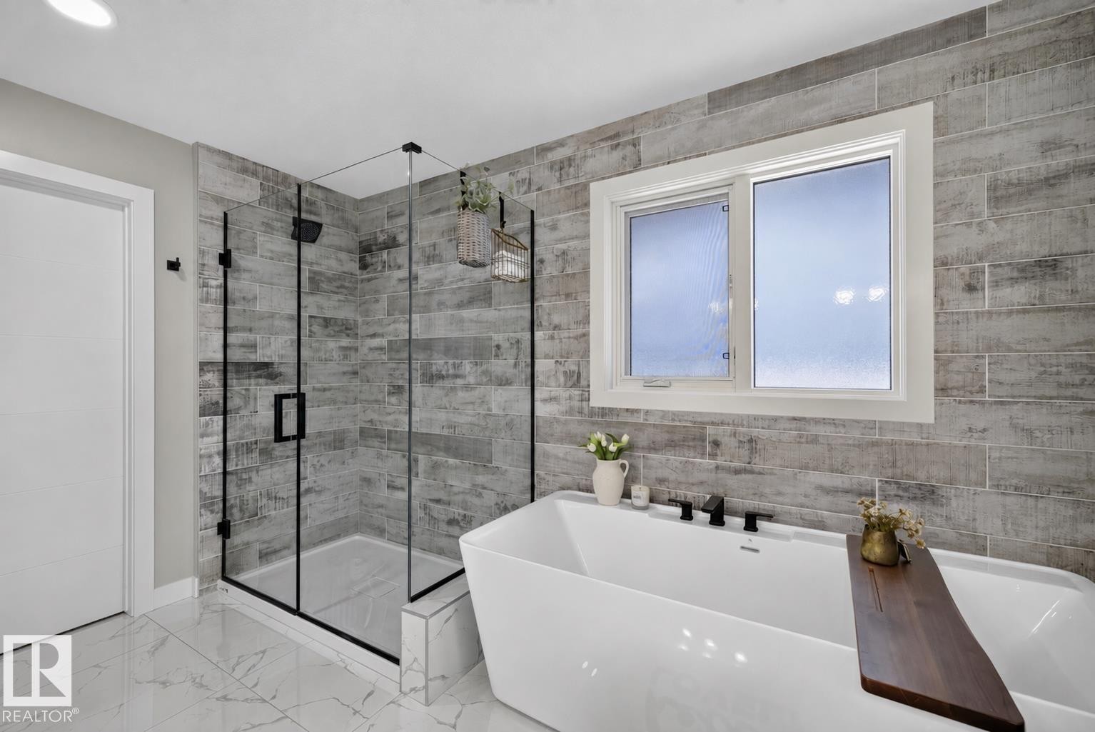 Contemporary bathroom featuring a freestanding soaking tub, a frameless glass shower enclosure with black hardware, white marble-look floor tiles, wood-look wall tiles, and a white-framed window - 1573 Esaiw Place Nw, Edmonton, AB
