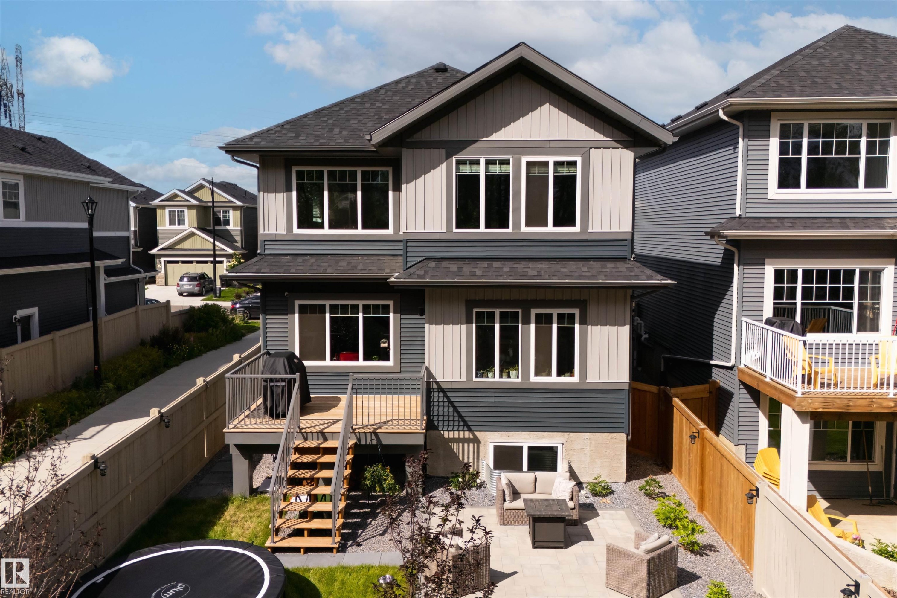 Two-story residence featuring multi-tone siding, a rear deck with stairs, a lower-level patio, a fenced yard, and multiple large windows - 1573 Esaiw Place Nw, Edmonton, AB
