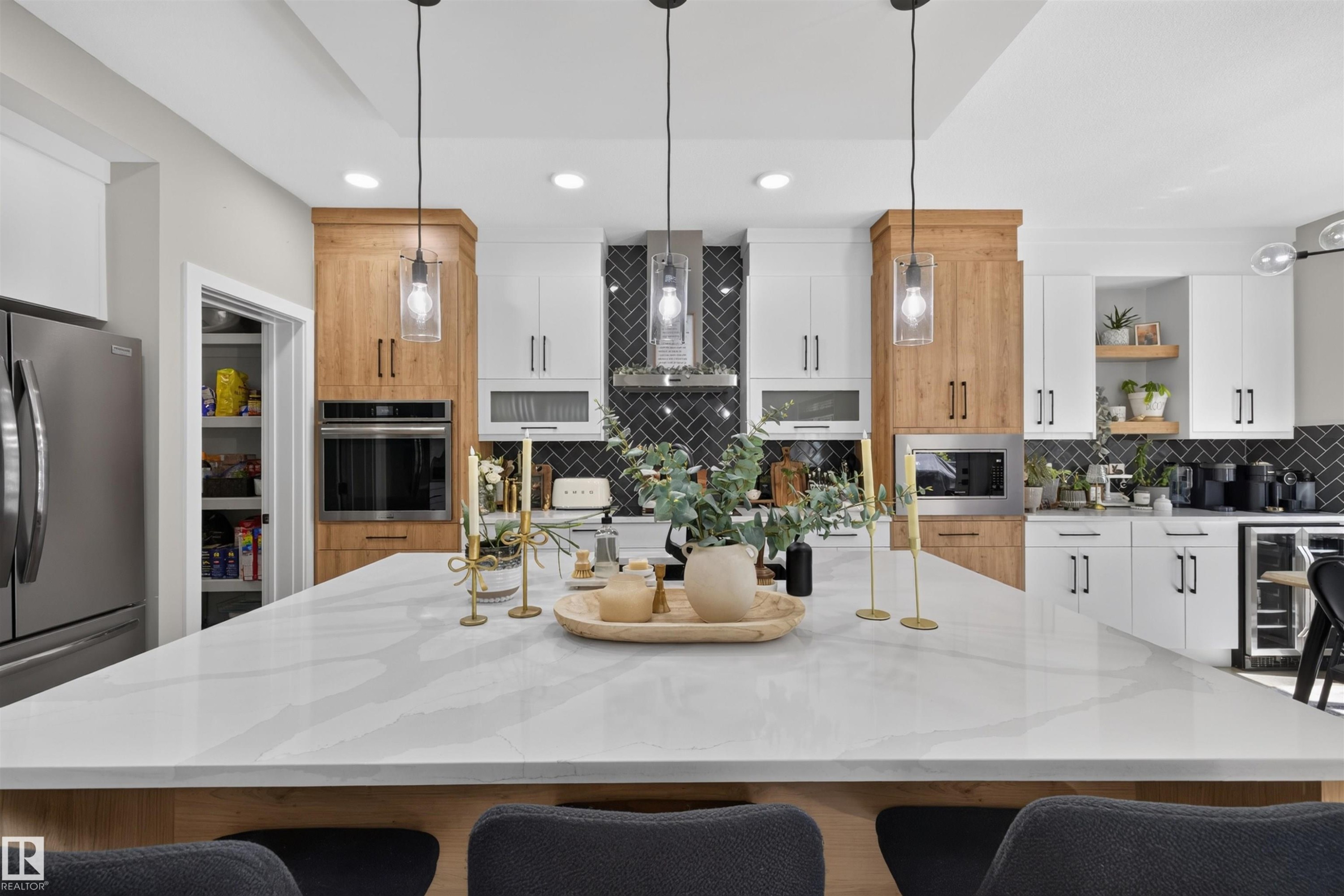 Spacious kitchen featuring a large central island with a light-toned countertop, contrasting white and wood-finish cabinetry, a dark herringbone tile backsplash, and built-in stainless steel appliances - 1573 Esaiw Place Nw, Edmonton, AB
