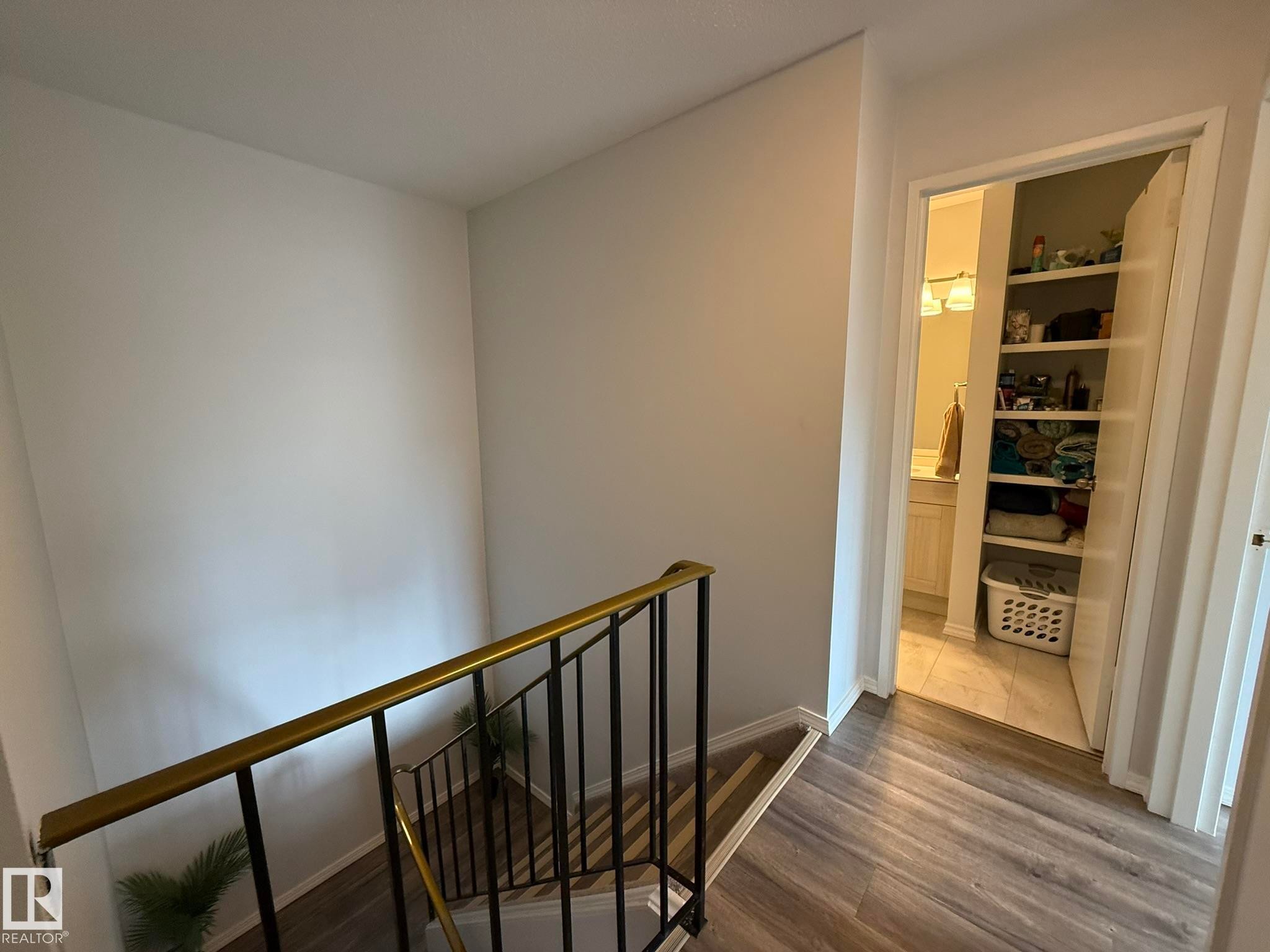 Upper-level landing featuring wood-finish flooring, a brass-finished handrail, and a storage closet with shelving - 166 Roseland Village, Edmonton, AB - Indoor Photo Showing Other Room