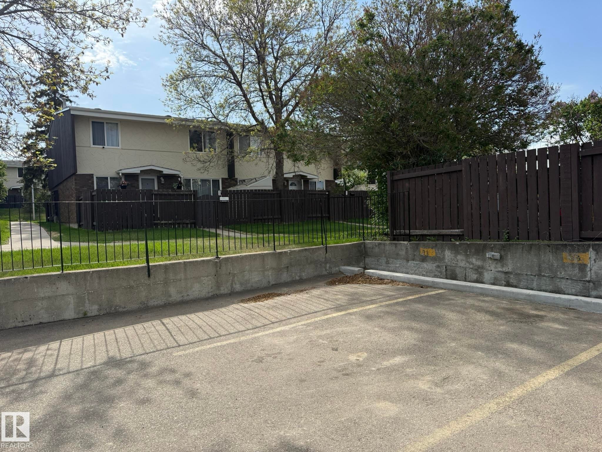 Paved parking area featuring a concrete retaining wall, a black metal fence, and a dark wood privacy fence - 166 Roseland Village, Edmonton, AB - Outdoor
