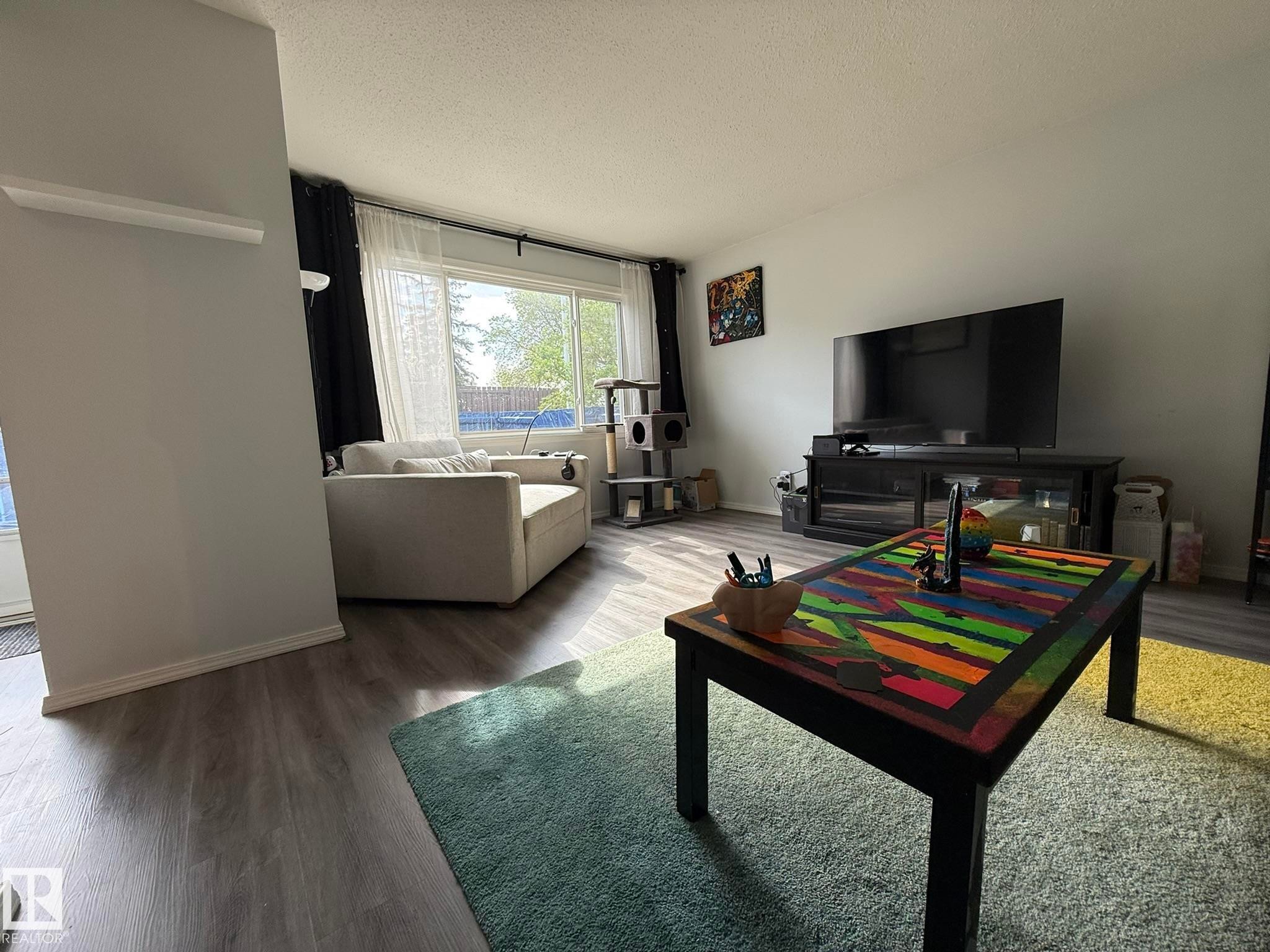 Living area featuring wood-finish flooring, light-toned walls, and a large window with white sheer and dark privacy drapes - 166 Roseland Village, Edmonton, AB - Indoor Photo Showing Living Room