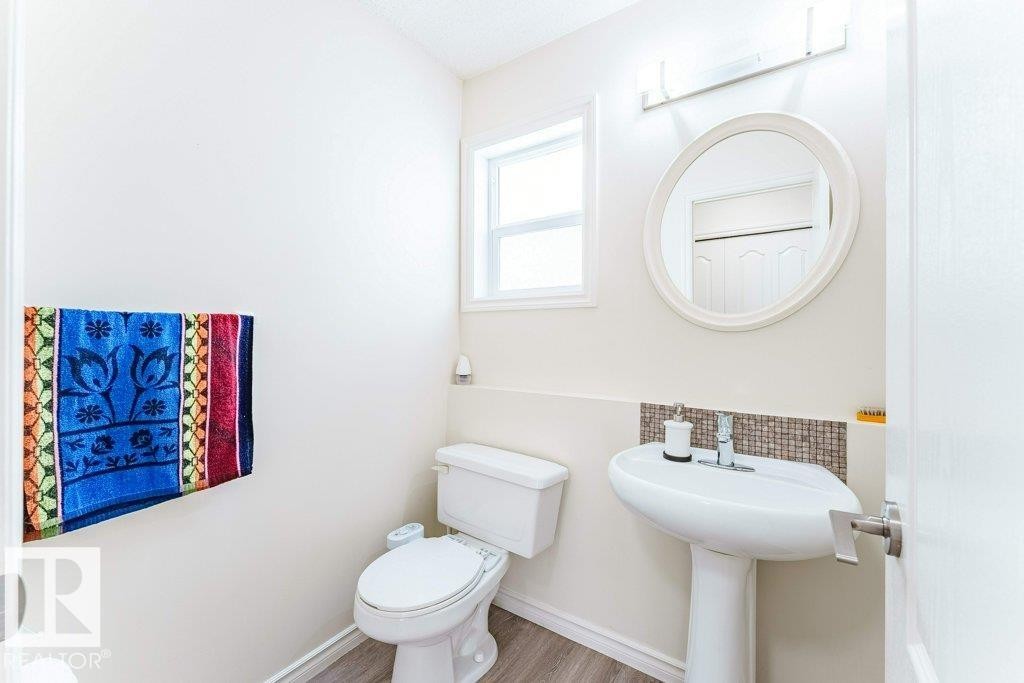 Bright powder room featuring a pedestal sink, round wall-mounted mirror, and wood-finish flooring - 3525 22 Street, Edmonton, AB - Indoor Photo Showing Bathroom