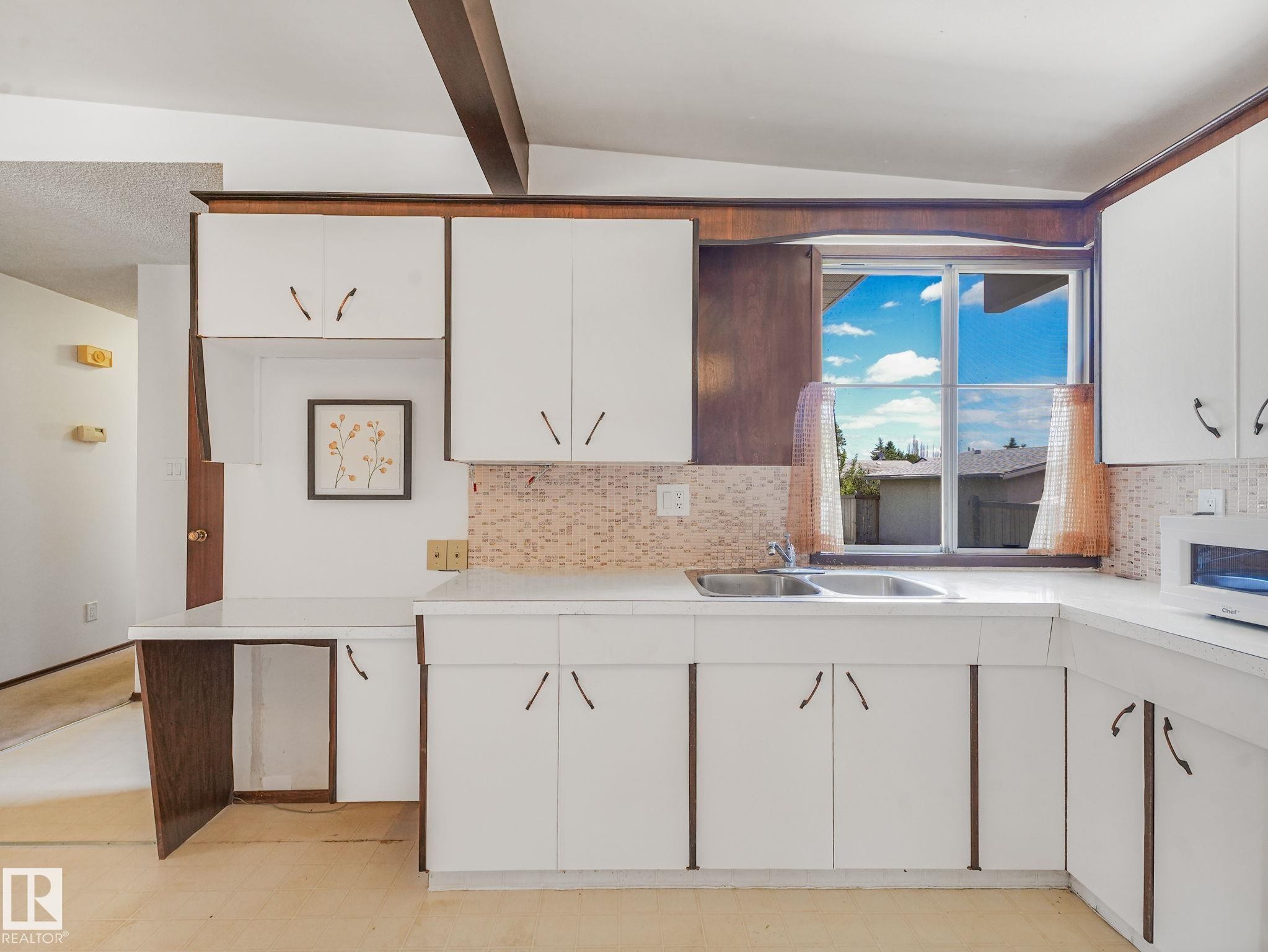 Kitchen featuring white cabinetry, a dual-basin stainless steel sink, patterned backsplash, a built-in desk area, and wood-finish ceiling beams - 15011 72A Street, Edmonton, AB