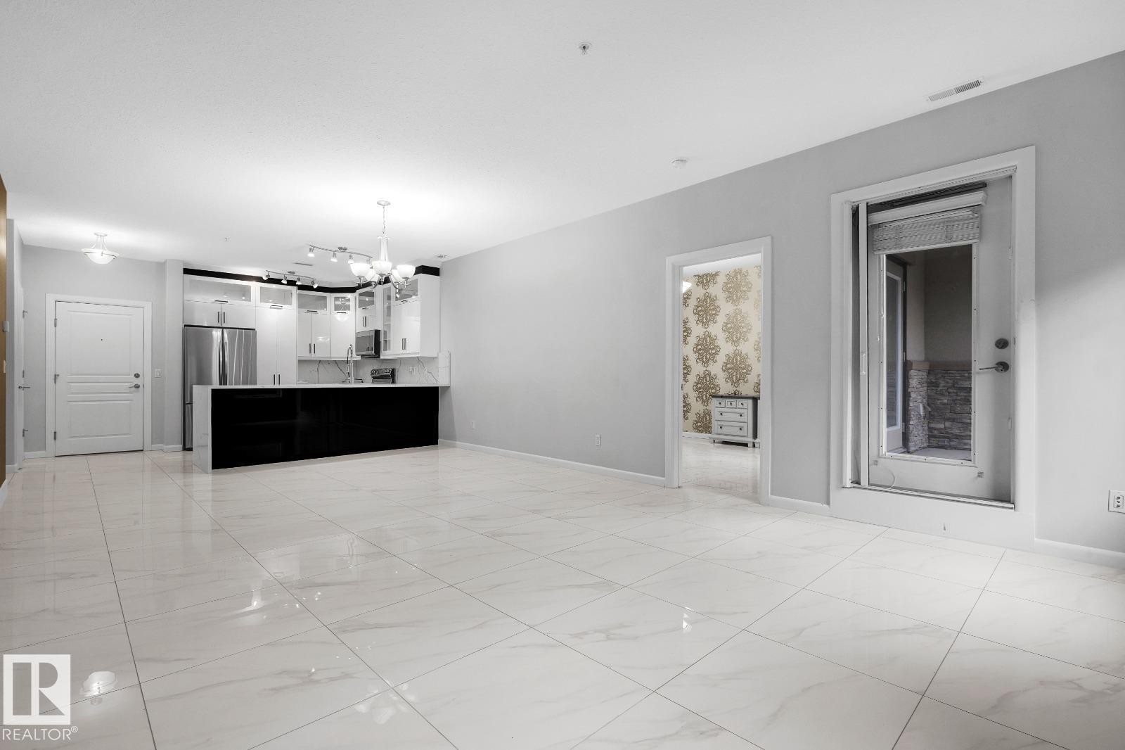 Open-concept living area featuring large format tile flooring and a kitchen with white cabinetry, stainless steel appliances, and a dark island - 105 160 Magrath Road, Edmonton, AB