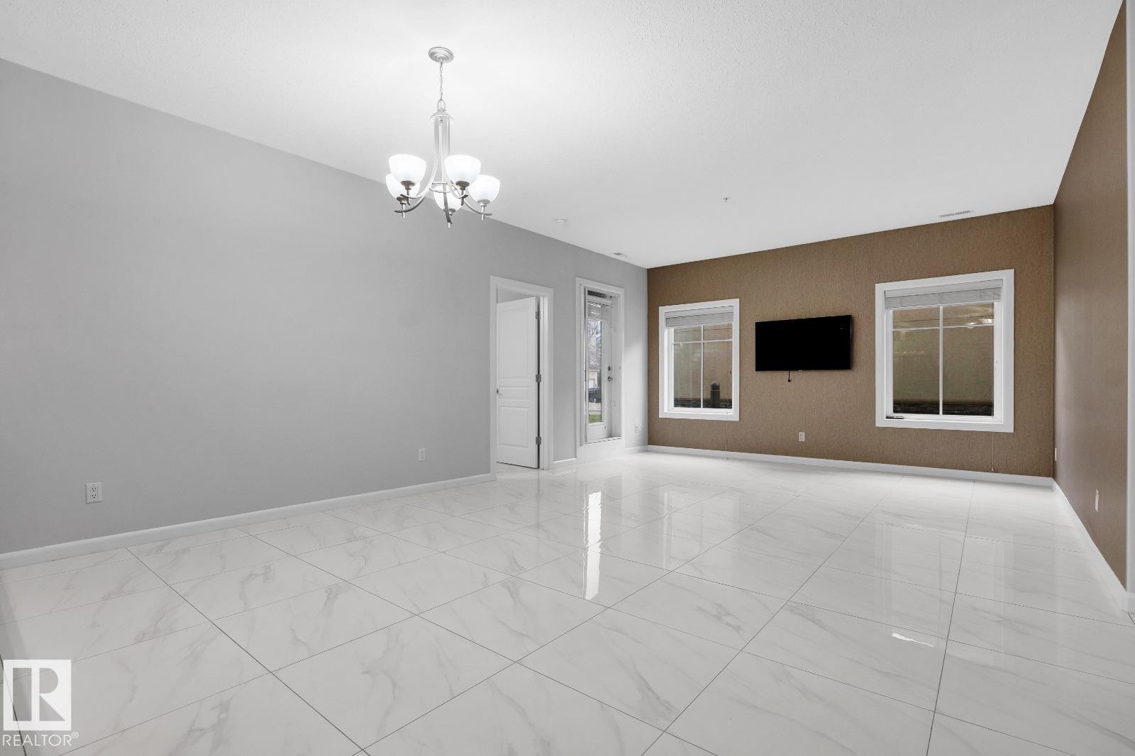 Spacious room featuring large format tile flooring, a contemporary chandelier, multiple windows, and a neutral color palette - 105 160 Magrath Road, Edmonton, AB
