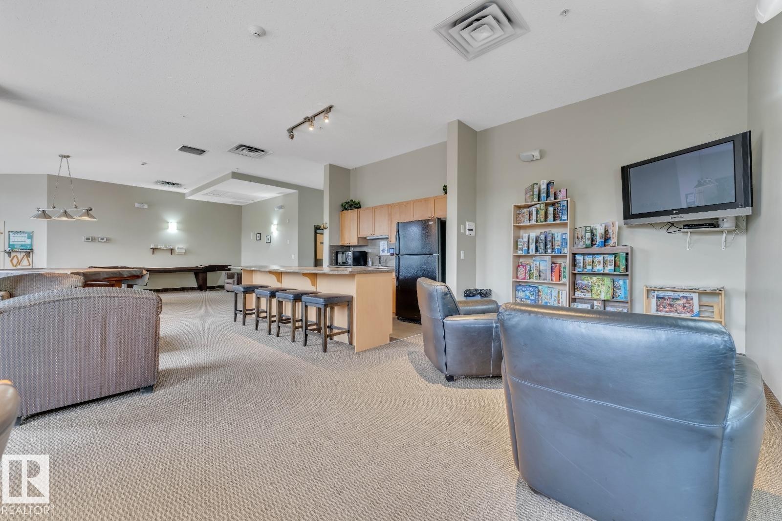 Spacious common area featuring textured carpeting, a kitchen area with light wood cabinetry, and a peninsula with bar seating - 105 160 Magrath Road, Edmonton, AB