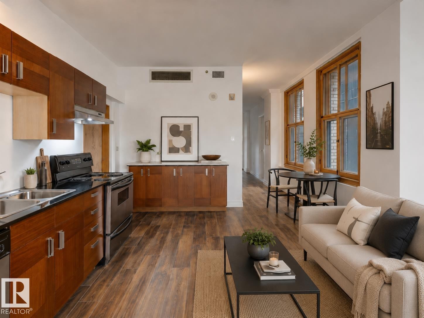Open-concept living space with wood-finish flooring, a kitchen featuring dark cabinetry and stainless steel appliances, and large multi-pane windows with natural wood frames - 210 10134 100 Street, Edmonton, AB