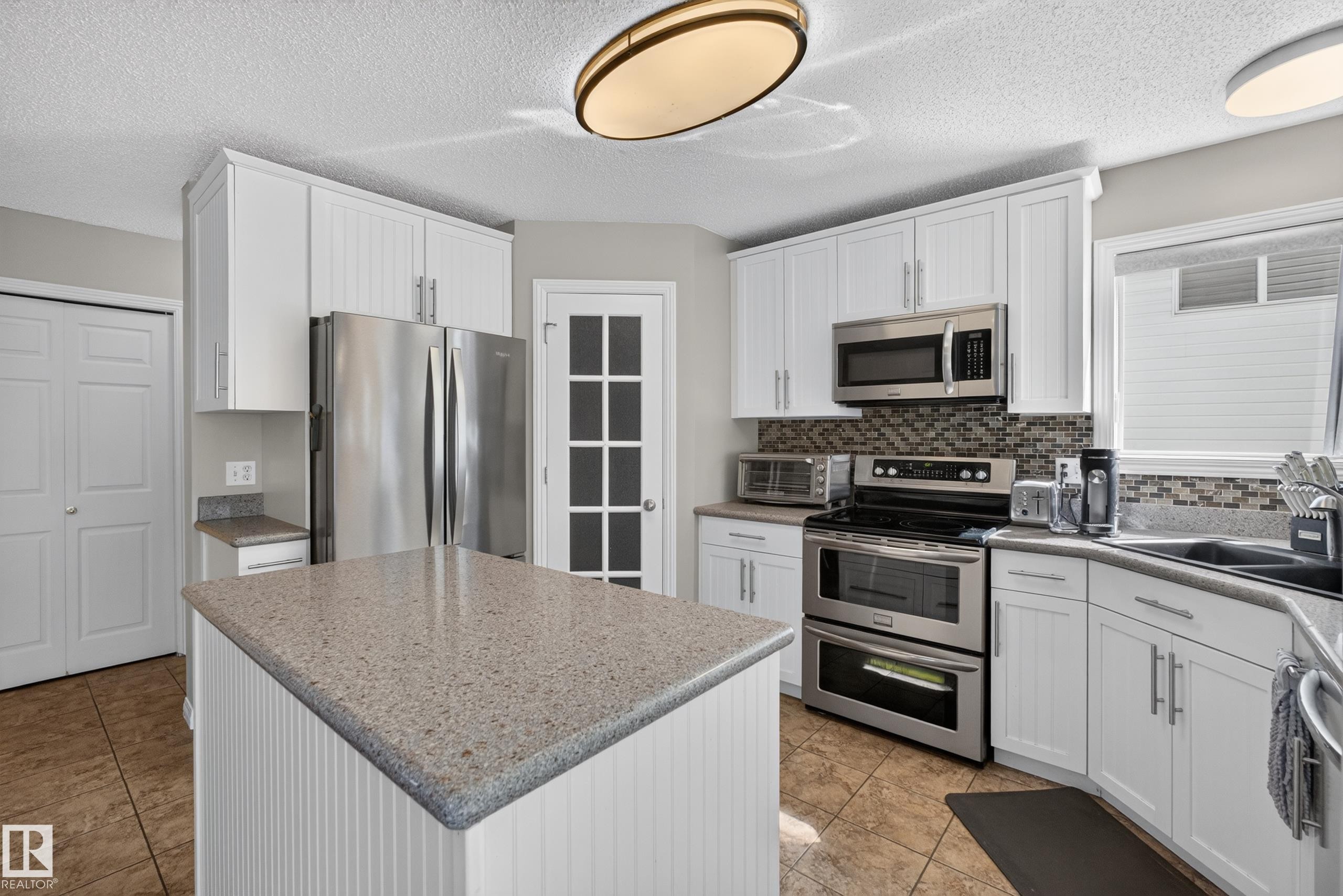 Kitchen featuring white cabinetry, a central island with a speckled countertop, stainless steel appliances, a mosaic tile backsplash, and tile flooring - 2327 37B Avenue, Edmonton, AB