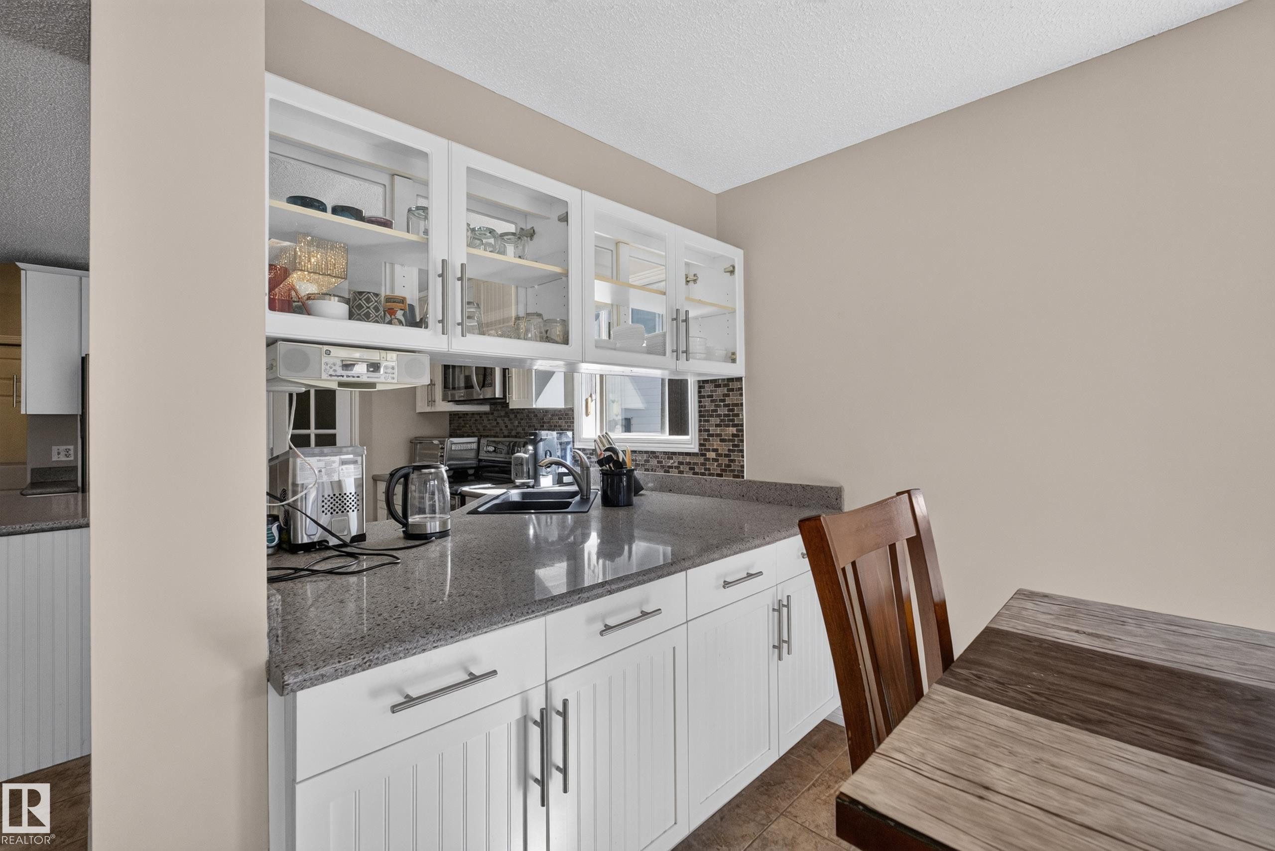 Kitchen featuring white shaker-style cabinetry with brushed nickel hardware, glass-front upper cabinets, speckled gray countertops, and a mosaic tile backsplash - 2327 37B Avenue, Edmonton, AB
