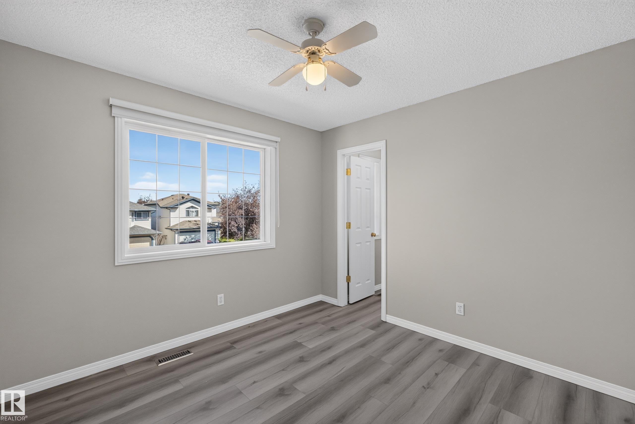 Bright interior room featuring a large window with white trim, wood-finish flooring, a ceiling fan with integrated lighting, a white painted door, and light gray wall paint - 2327 37B Avenue, Edmonton, AB