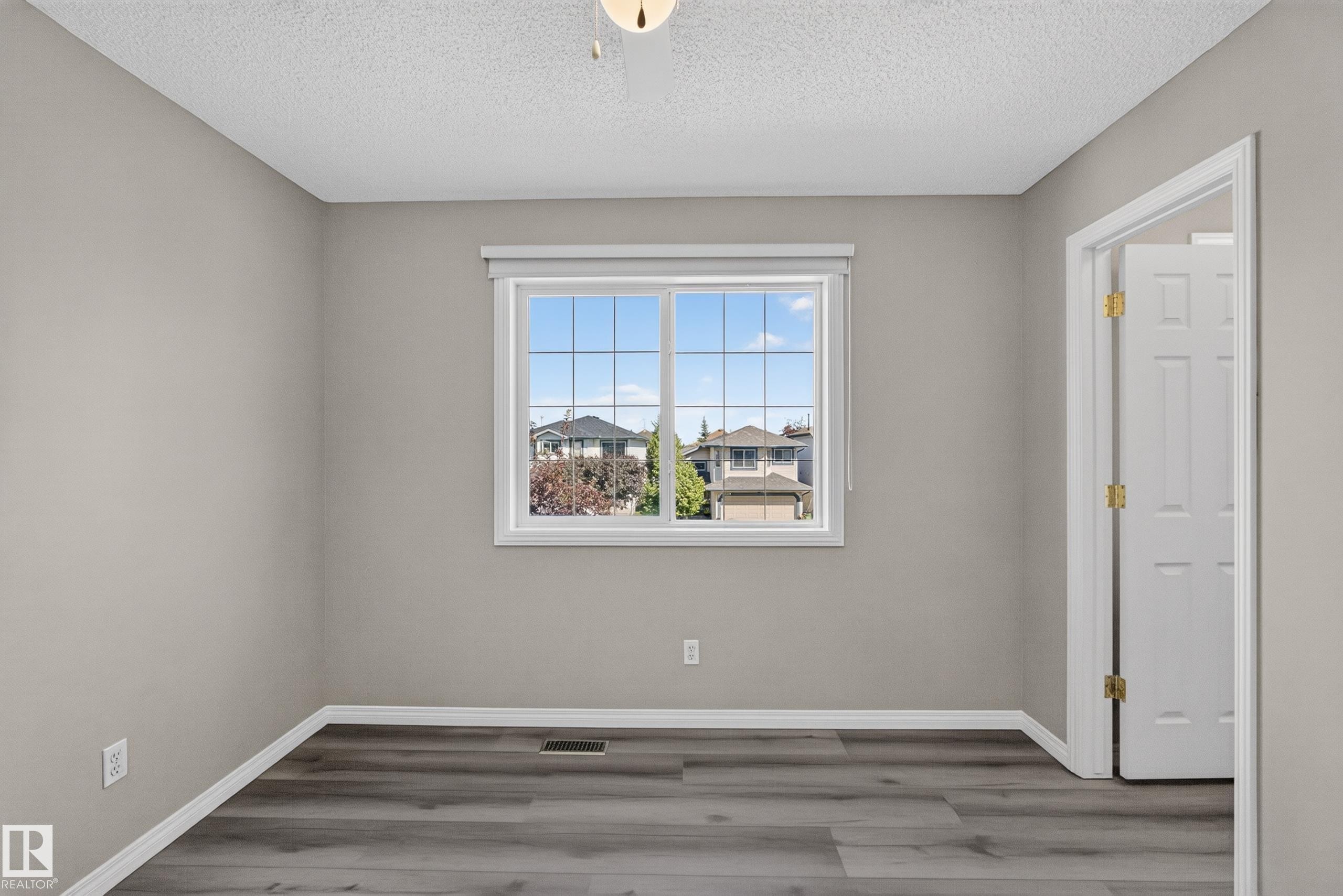 Spacious room featuring wood-finish flooring, a white trim window, neutral wall paint, and a white paneled door with brass hardware - 2327 37B Avenue, Edmonton, AB