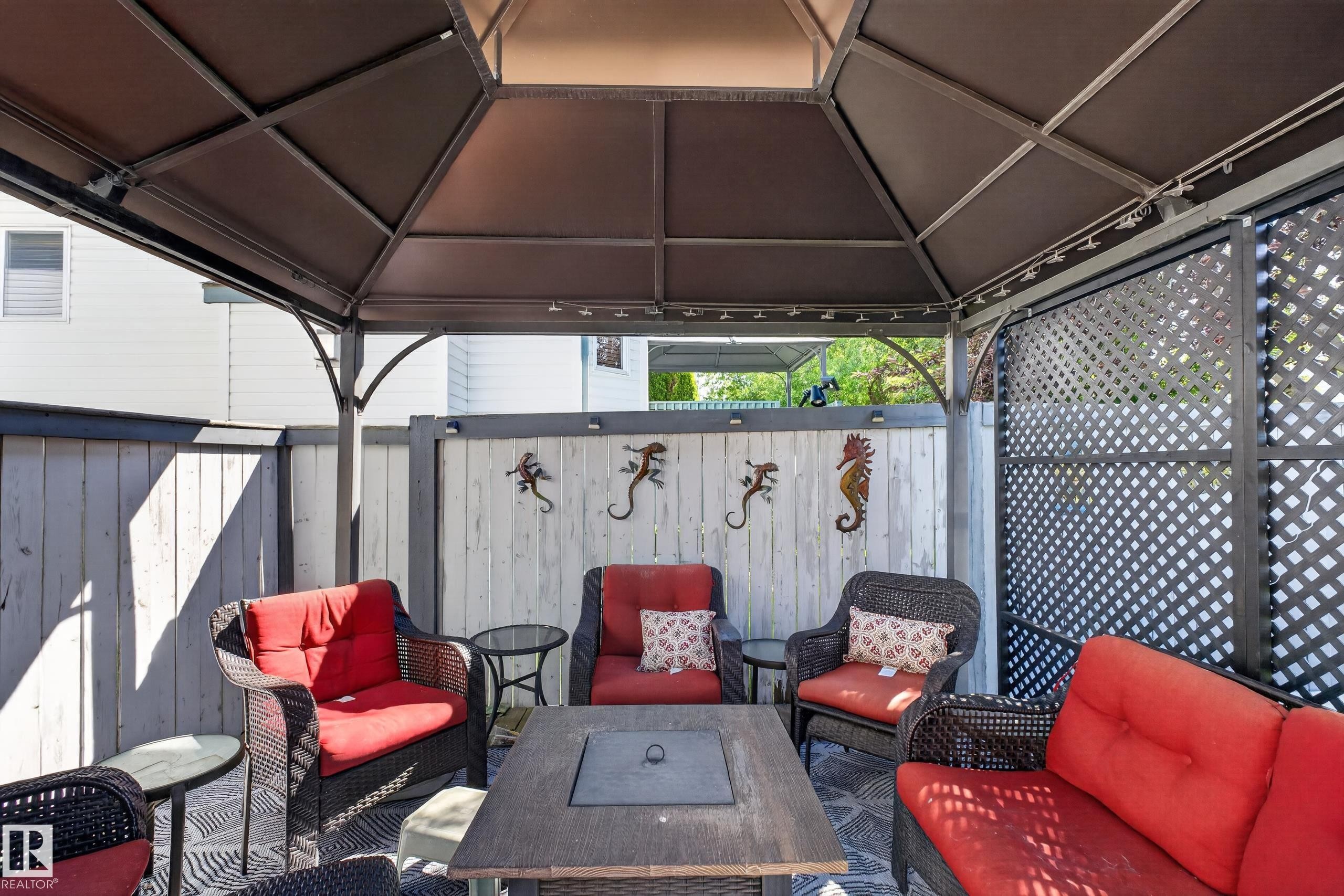 Covered outdoor patio area featuring a dark-framed gazebo structure with overhead lighting, a built-in fire pit table, light-toned wood fencing, and dark lattice privacy screens - 2327 37B Avenue, Edmonton, AB