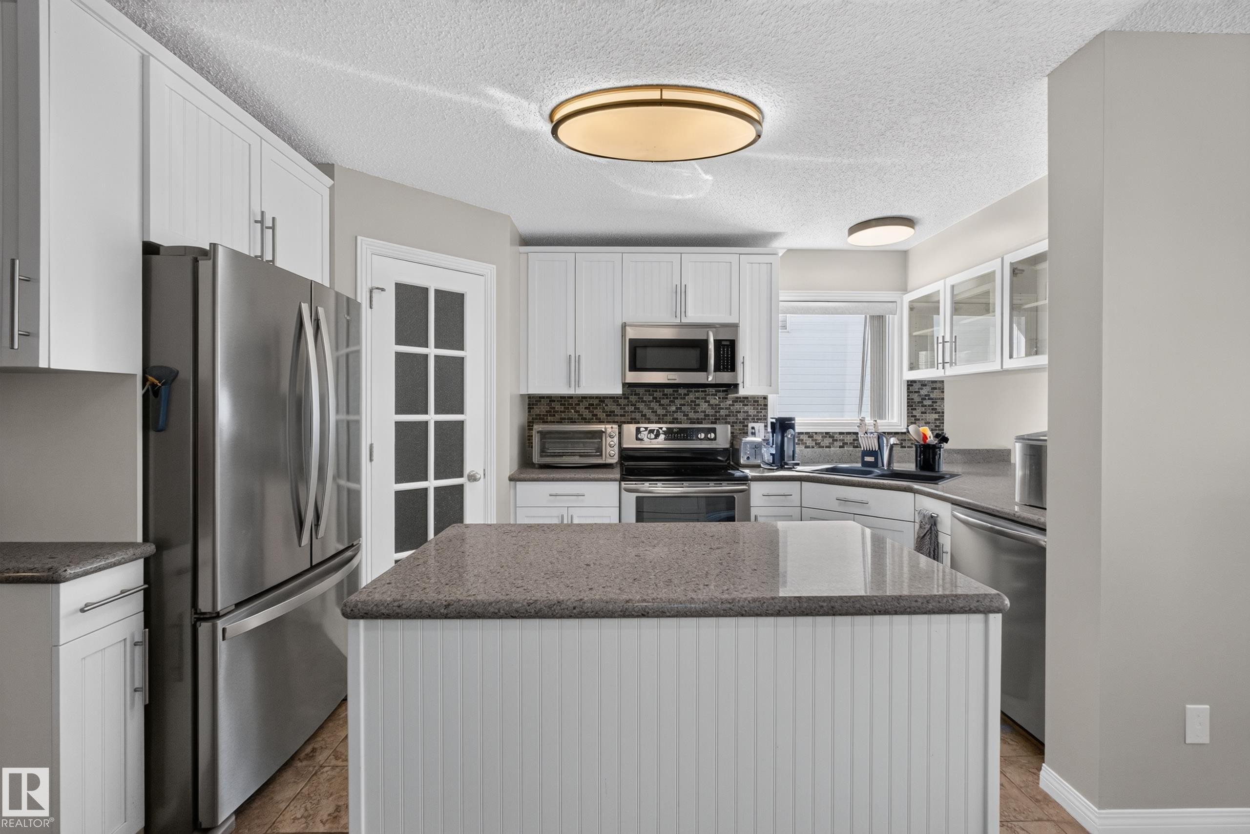 Kitchen featuring a central island with beadboard paneling, light-toned countertops, white cabinetry, stainless steel appliances, and a mosaic tile backsplash - 2327 37B Avenue, Edmonton, AB