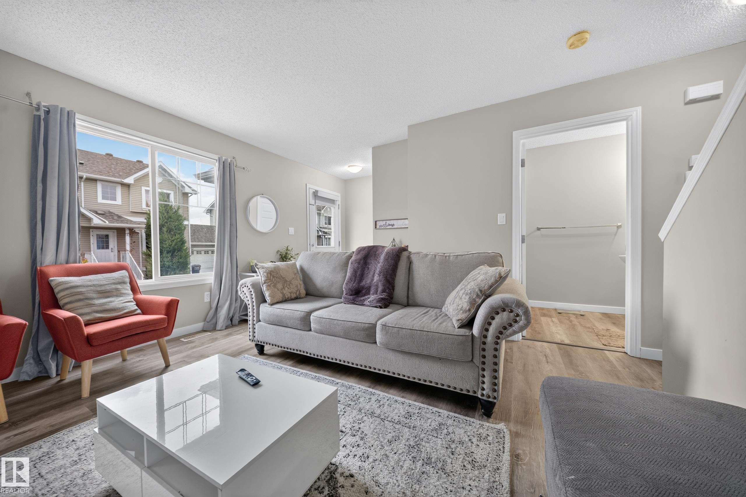 Spacious living area featuring wood-finish flooring, light grey wall paint, a large picture window, and visible staircase - 59 2503 24 Street, Edmonton, AB