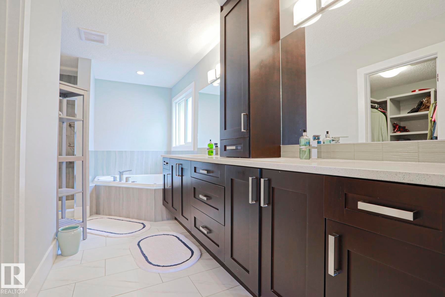 Bathroom featuring a corner soaking tub, a dual vanity with dark wood cabinetry and light countertops, and a full-width mirror - 1304 Ainslie Wynd Sw, Edmonton, AB - Indoor Photo Showing Bathroom