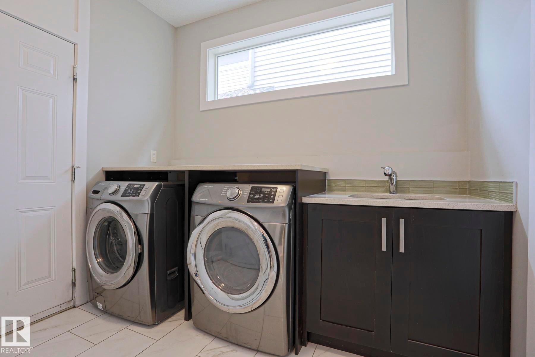 Laundry area featuring a utility sink, dark cabinetry with silver hardware, light-toned countertops, and a horizontal window - 1304 Ainslie Wynd Sw, Edmonton, AB - Indoor Photo Showing Laundry Room
