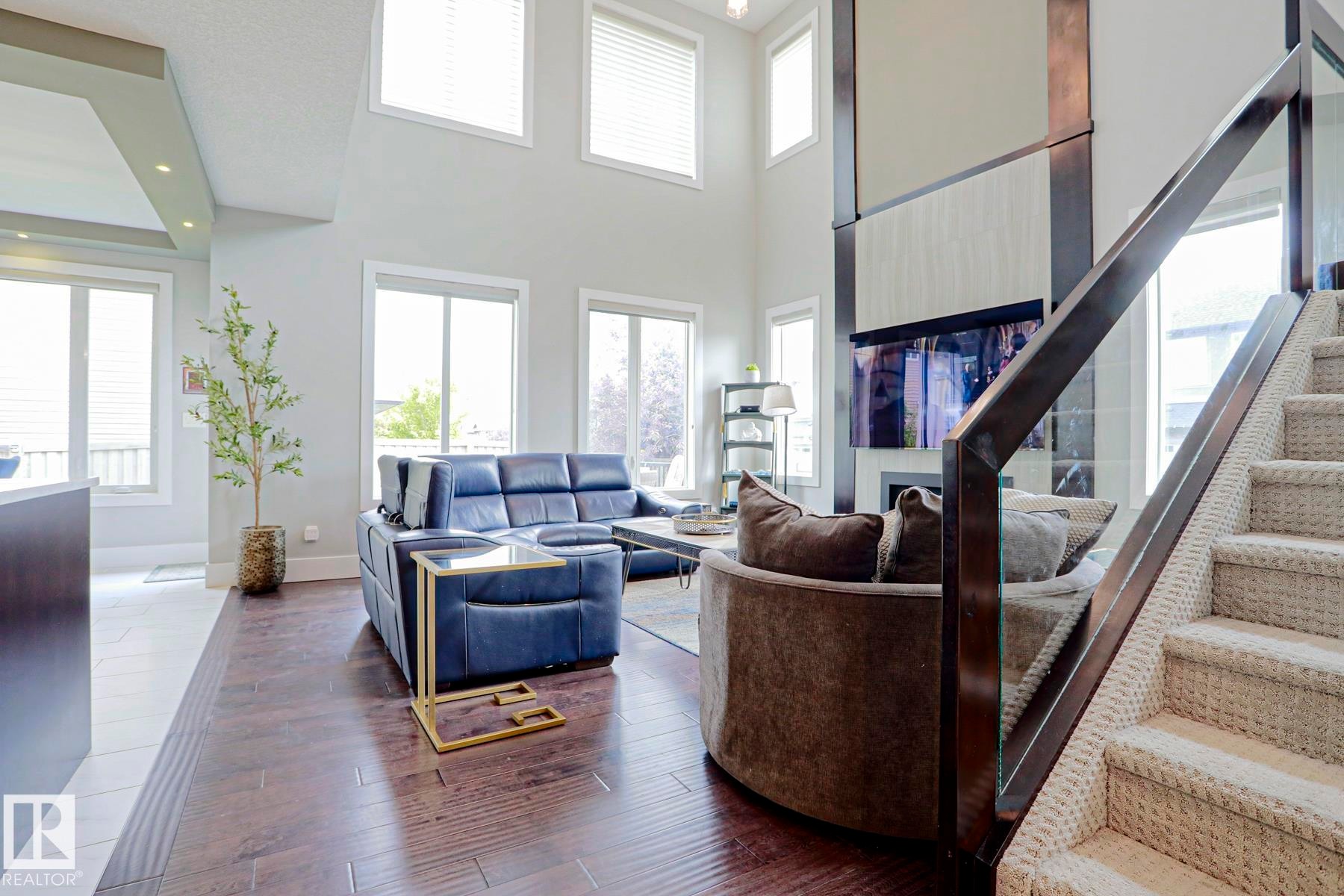 Two-story living area featuring wood-finish flooring, a contemporary fireplace with mounted television, and a carpeted staircase with glass railing - 1304 Ainslie Wynd Sw, Edmonton, AB - Indoor Photo Showing Other Room