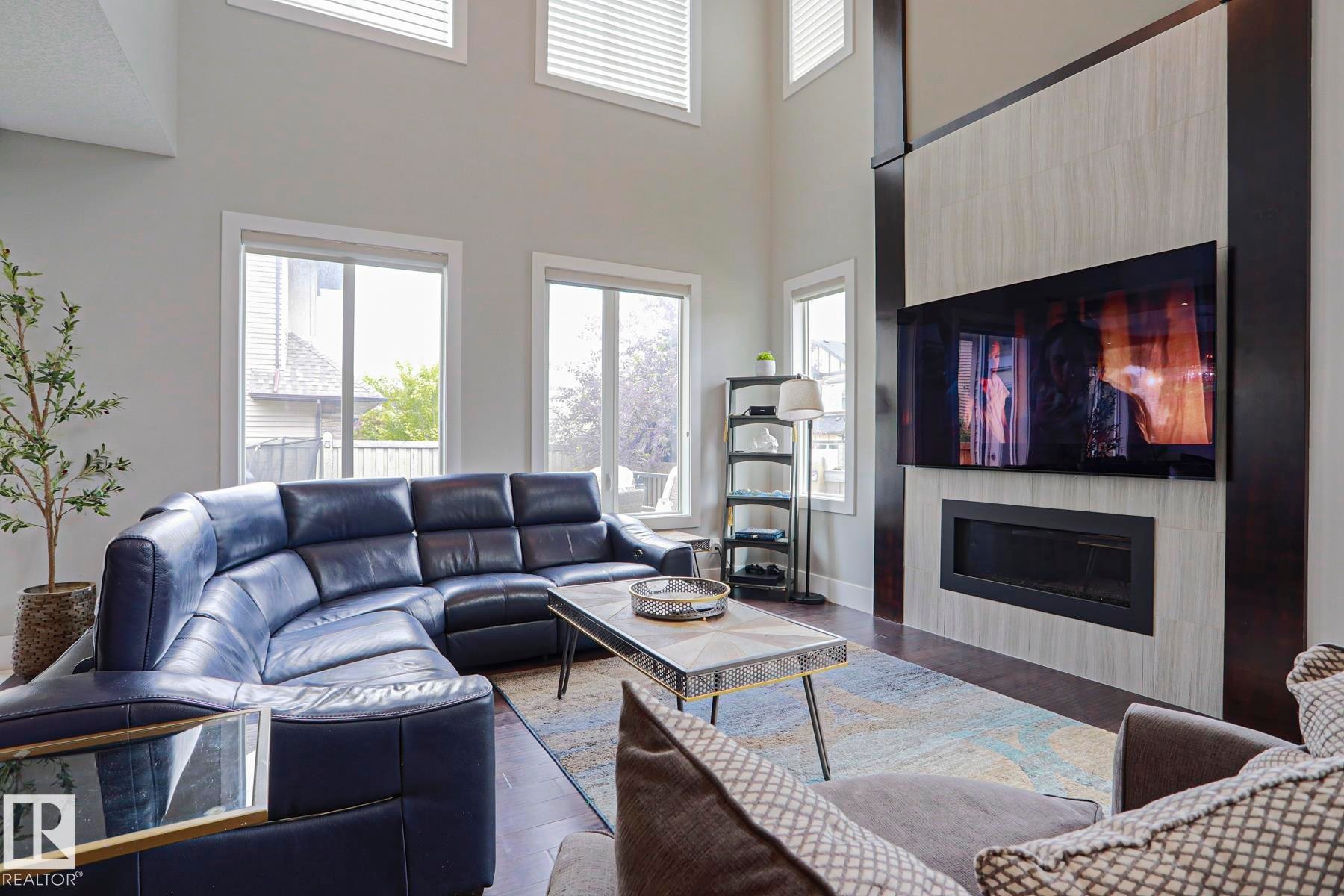 Living space featuring a two-story ceiling, multiple windows with blinds, wood-finish flooring, a built-in electric fireplace, and a large flat-screen display - 1304 Ainslie Wynd Sw, Edmonton, AB - Indoor Photo Showing Living Room With Fireplace