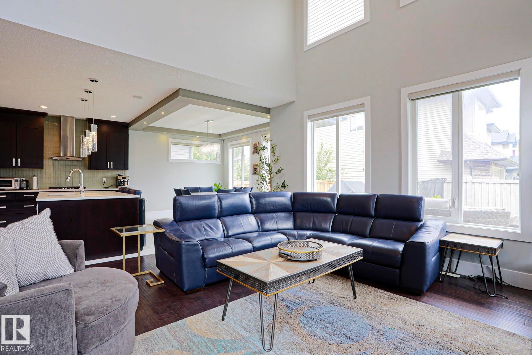 Open-concept living space featuring wood-finish flooring, a two-story ceiling, multiple windows, a kitchen island with seating, and dark cabinetry - 1304 Ainslie Wynd Sw, Edmonton, AB - Indoor Photo Showing Living Room