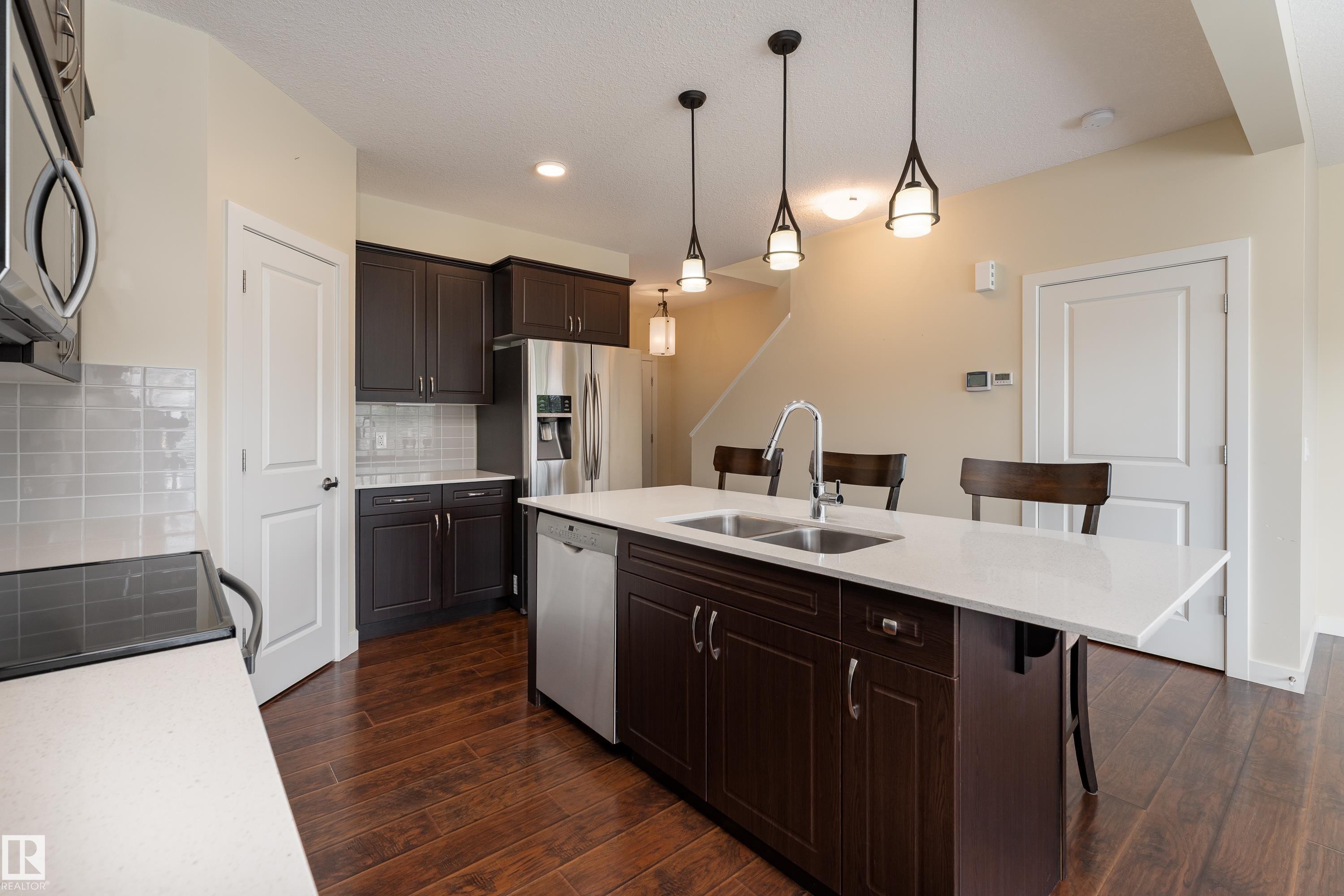 Kitchen featuring dark wood cabinetry, stainless steel appliances, a center island with double sink, white countertops, and wood-finish flooring - 8589 Cushing Place Sw, Edmonton, AB - Indoor Photo Showing Kitchen With Stainless Steel Kitchen With Double Sink With Upgraded Kitchen