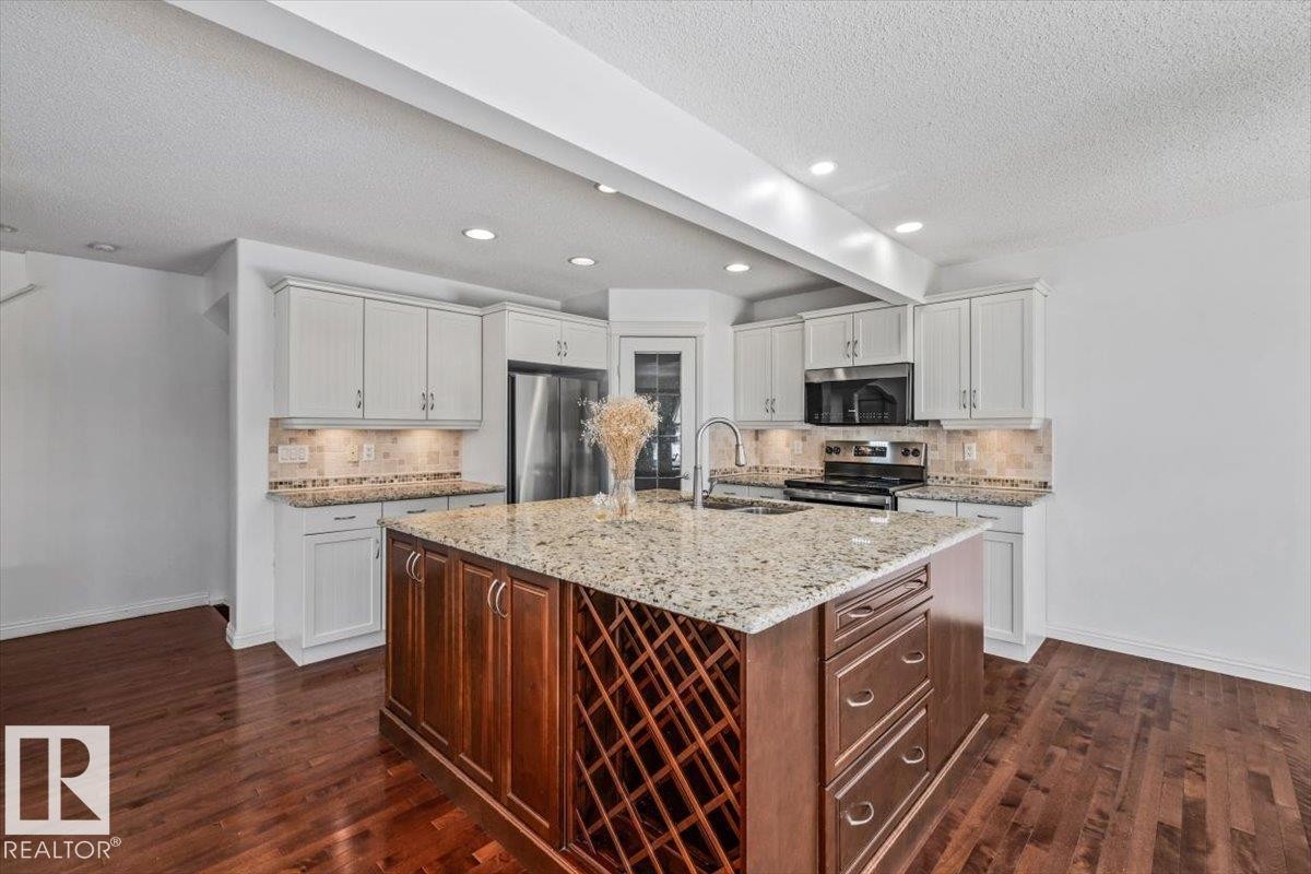 Kitchen featuring a spacious island with integrated wine storage, light-toned perimeter cabinetry, stainless steel appliances, a tile backsplash, and wood-finish flooring - 2407 Hagen Way, Edmonton, AB