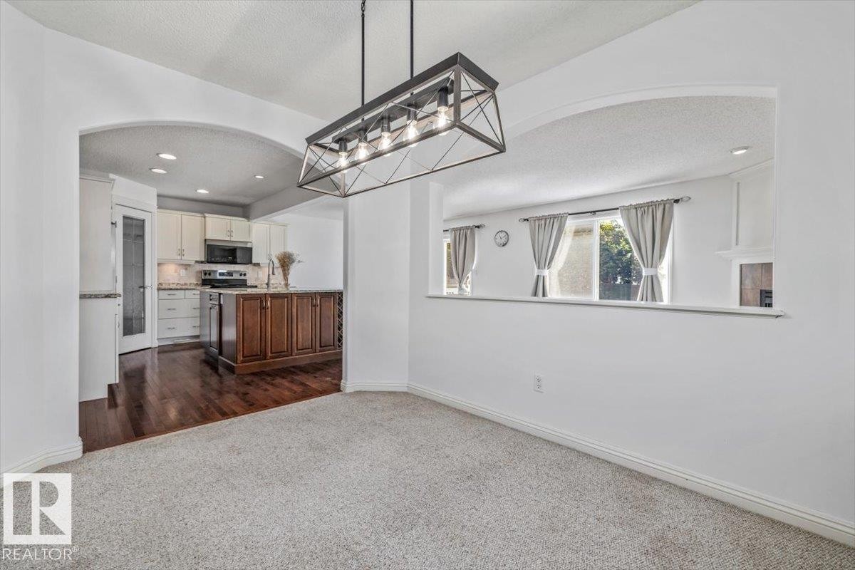 Carpeted dining area with a linear chandelier, arched passthrough, and a half wall opening to a kitchen with wood-finish flooring, a kitchen island, and white cabinetry - 2407 Hagen Way, Edmonton, AB