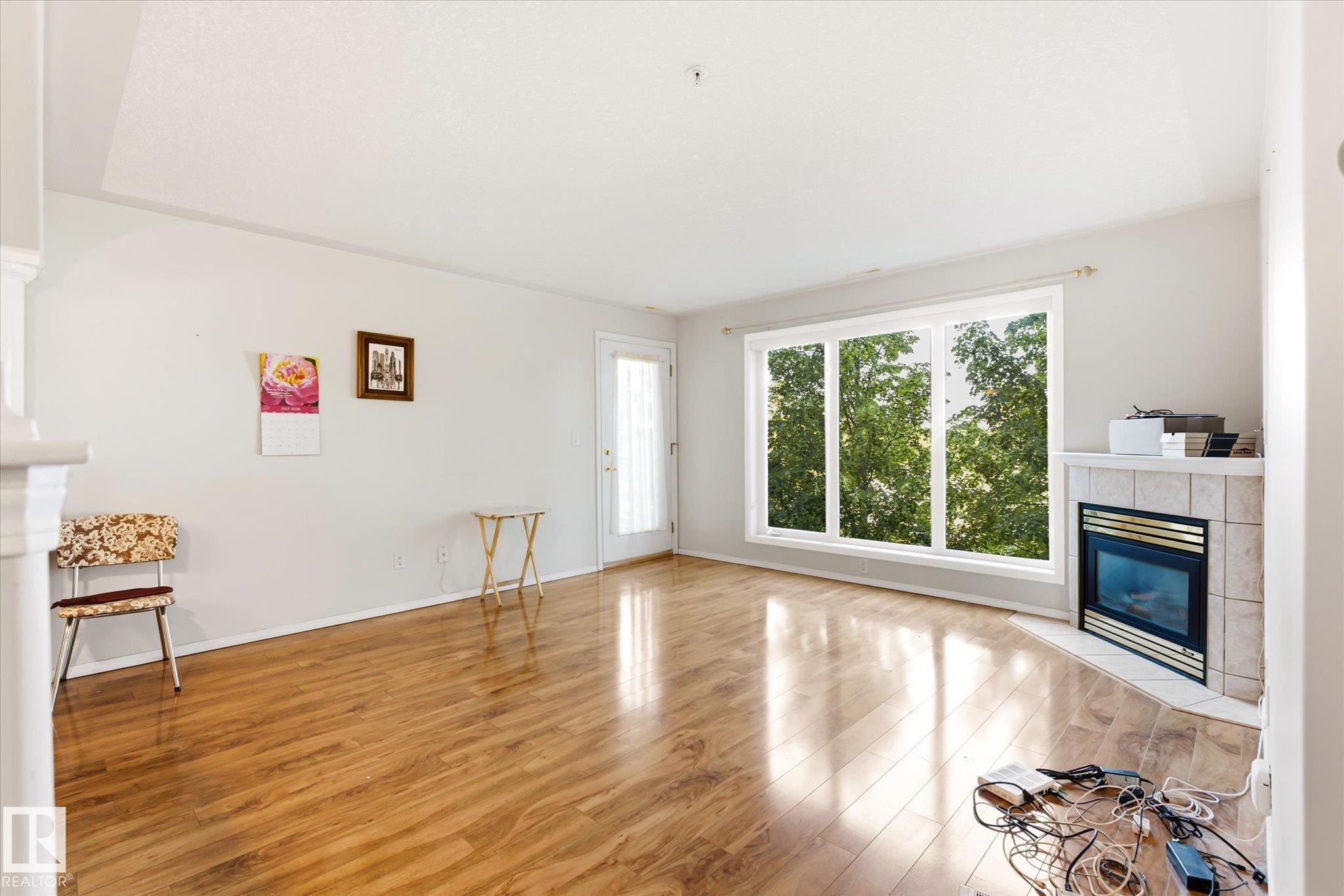 Spacious living area featuring wood-finish flooring, a tiled corner fireplace, a large picture window, and a glass-paneled door - #310 237 Youville Dr E Nw, Edmonton, AB - Indoor Photo Showing Other Room With Fireplace