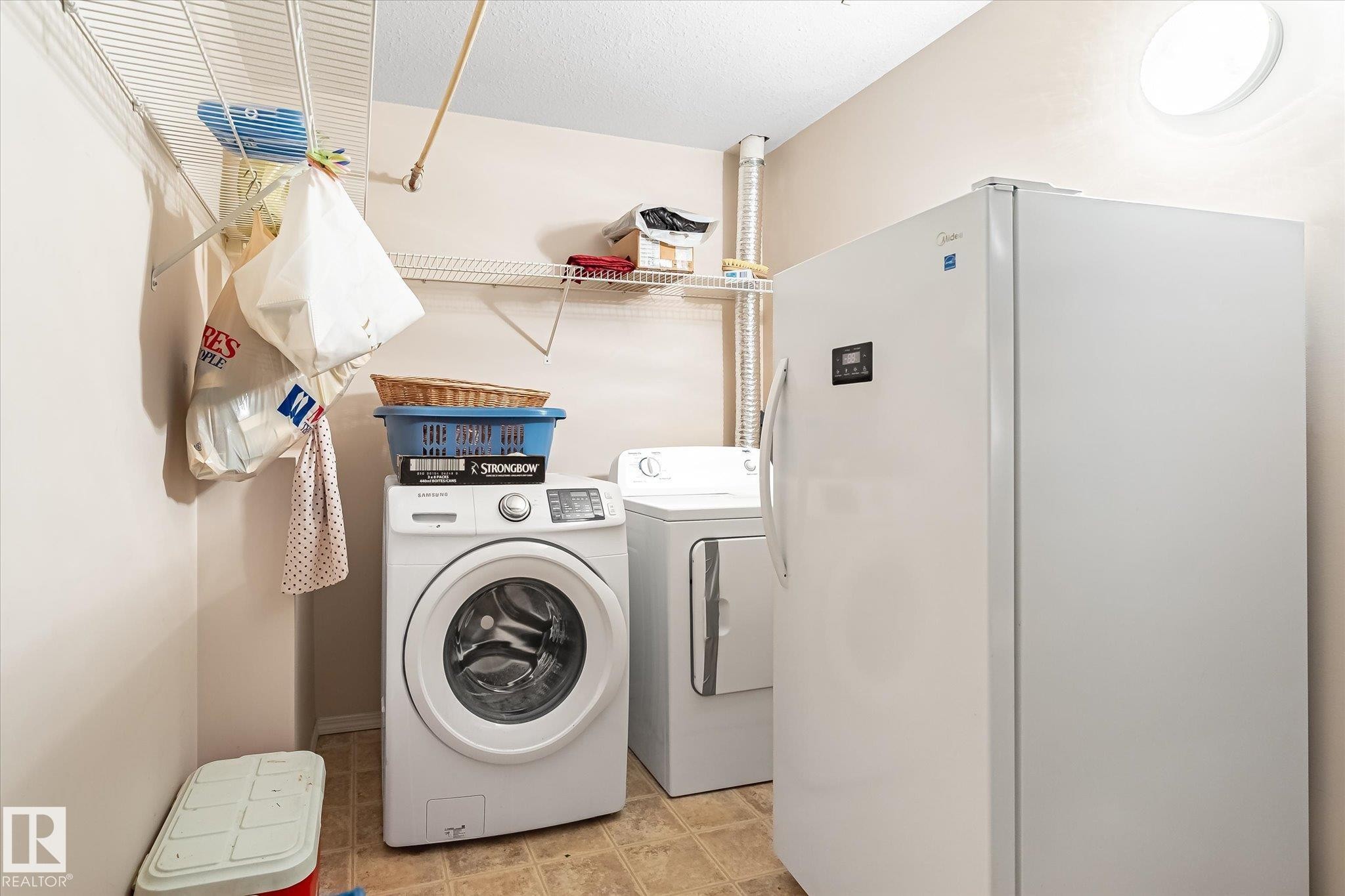 Utility area featuring a washer and dryer, additional freestanding refrigerator, overhead circular lighting, and wall-mounted wire shelving - #310 237 Youville Dr E Nw, Edmonton, AB - Indoor Photo Showing Laundry Room