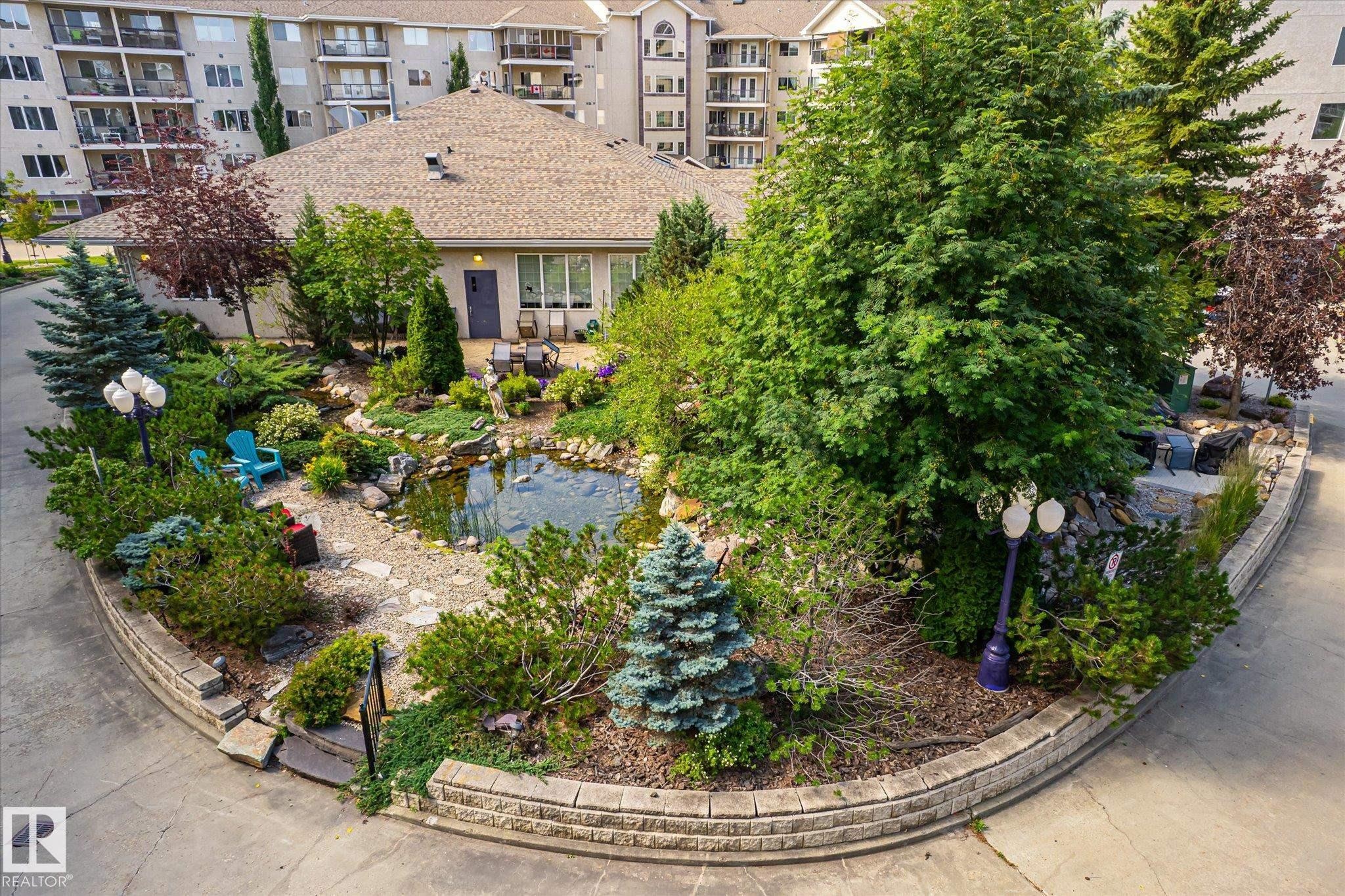 Lush landscaping featuring a central pond with rock accents, surrounded by varied evergreen and deciduous plantings - #310 237 Youville Dr E Nw, Edmonton, AB - Outdoor
