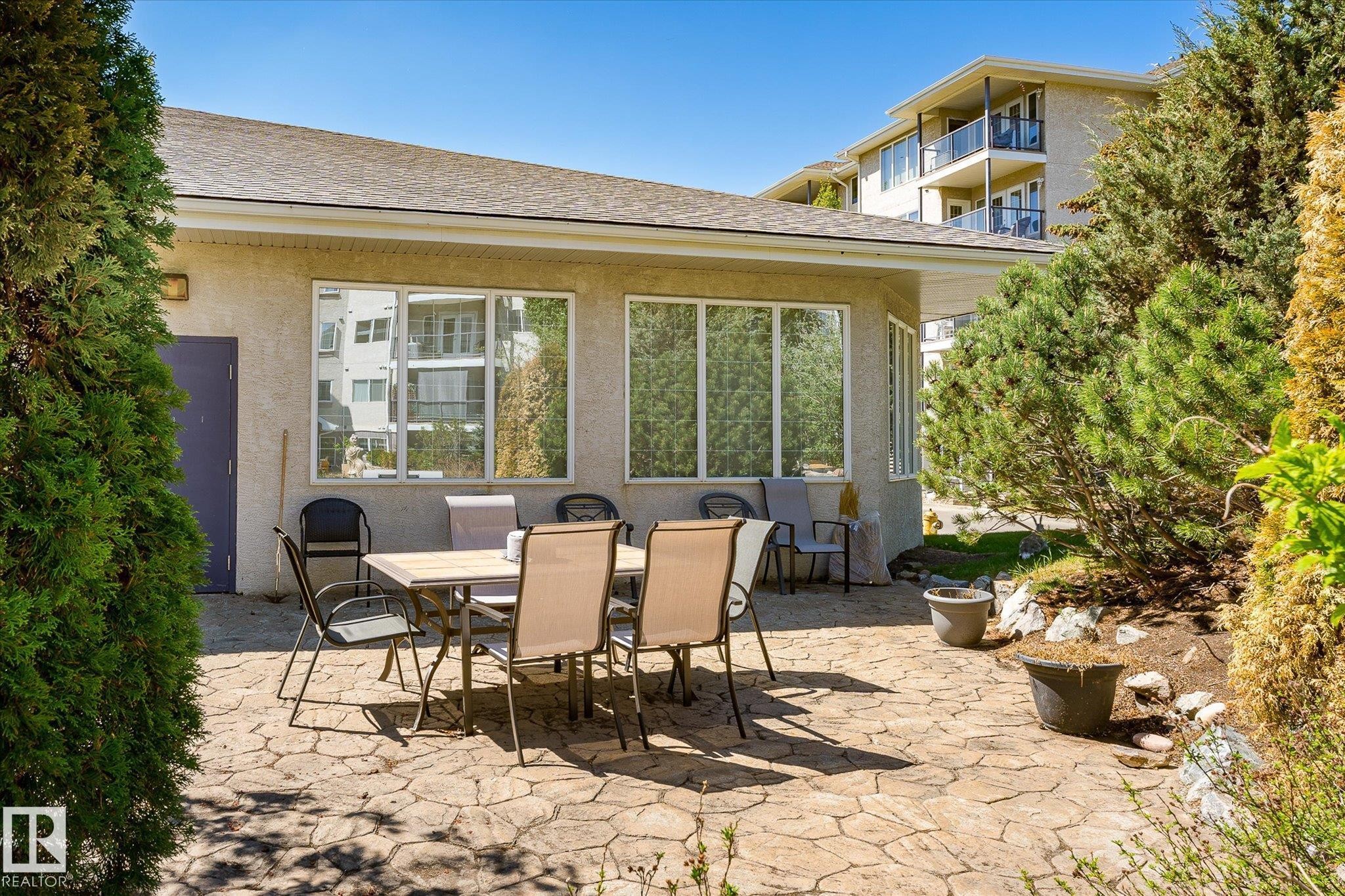 Stucco exterior with multiple large windows, a patterned concrete patio, and mature landscaping - #310 237 Youville Dr E Nw, Edmonton, AB - Outdoor With Deck Patio Veranda